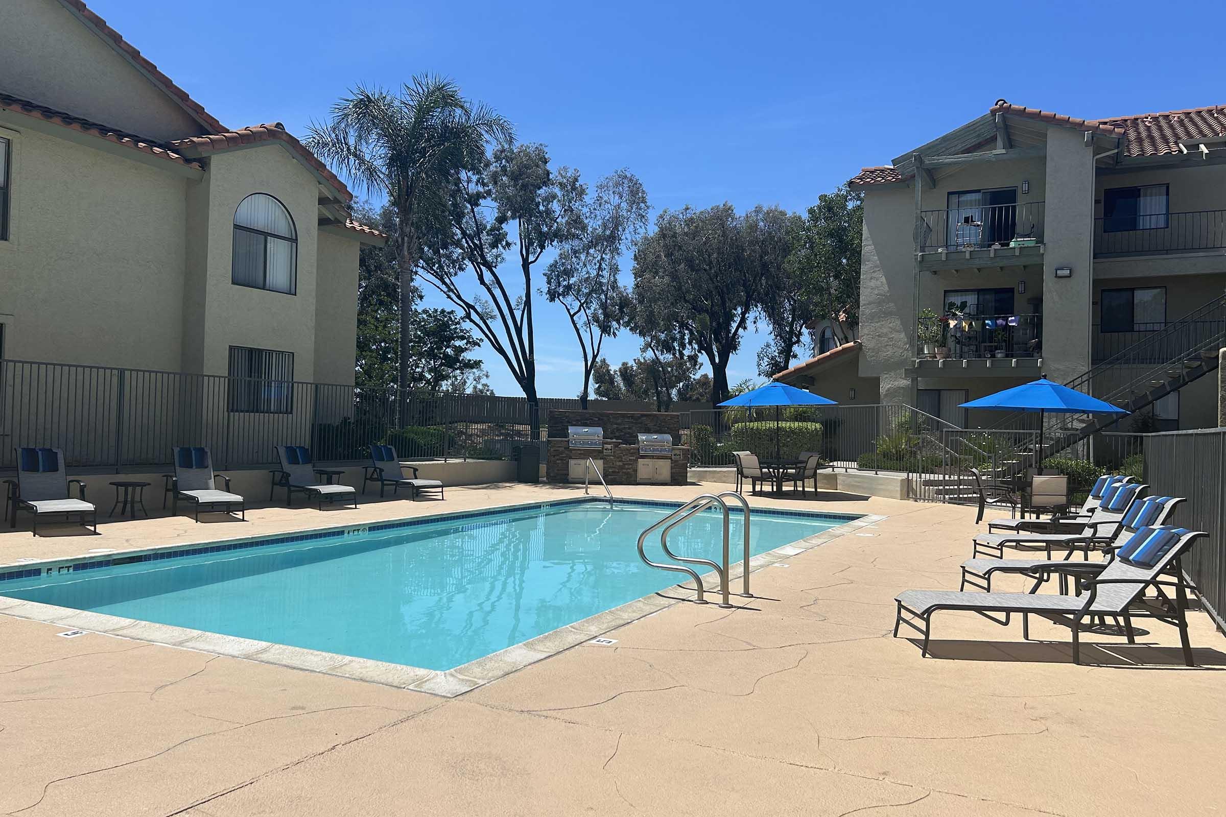 A sunny outdoor pool area surrounded by lounge chairs and umbrellas. The pool is clear and inviting, bordered by a fence. Nearby, there are trees and two multi-story buildings, indicating a residential complex. The overall atmosphere is relaxed and perfect for leisure activities.
