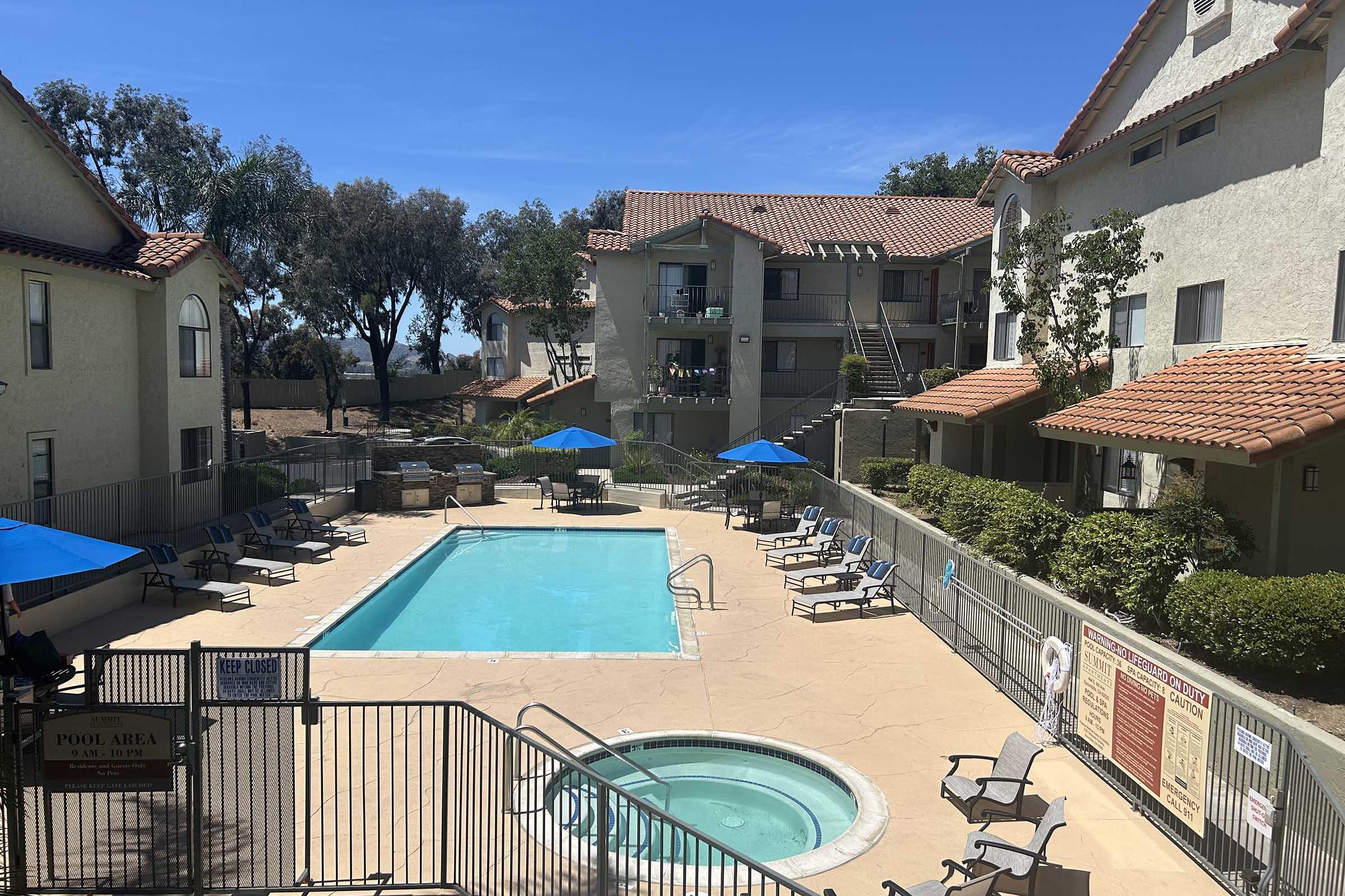 View of an outdoor pool area in an apartment complex. The pool is surrounded by lounge chairs and blue umbrellas, with a hot tub nearby. Two buildings with balconies and greenery are visible in the background under a clear blue sky.