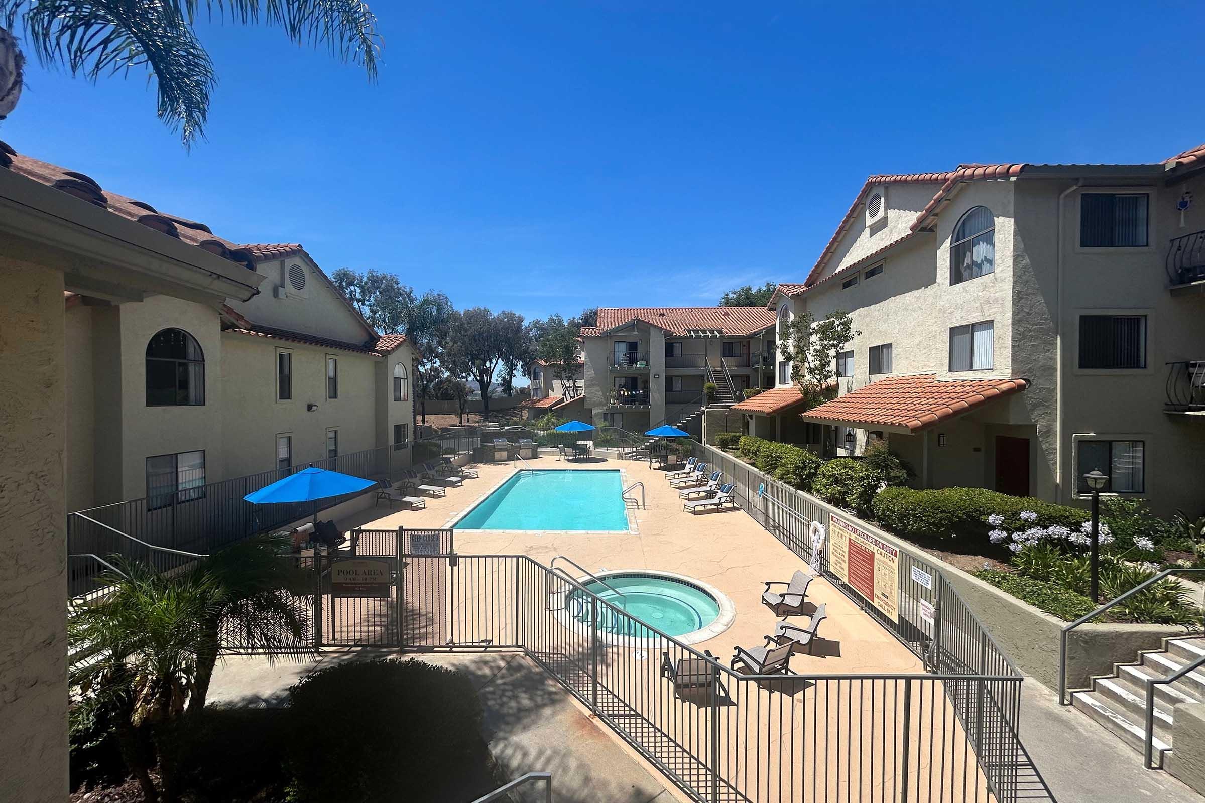 A sunny residential area featuring a pool surrounded by lounge chairs and umbrellas. The view includes multiple buildings with red-tiled roofs and palm trees, creating a relaxed atmosphere. The scene is clear and vibrant, highlighting the blue sky and well-maintained landscaping.
