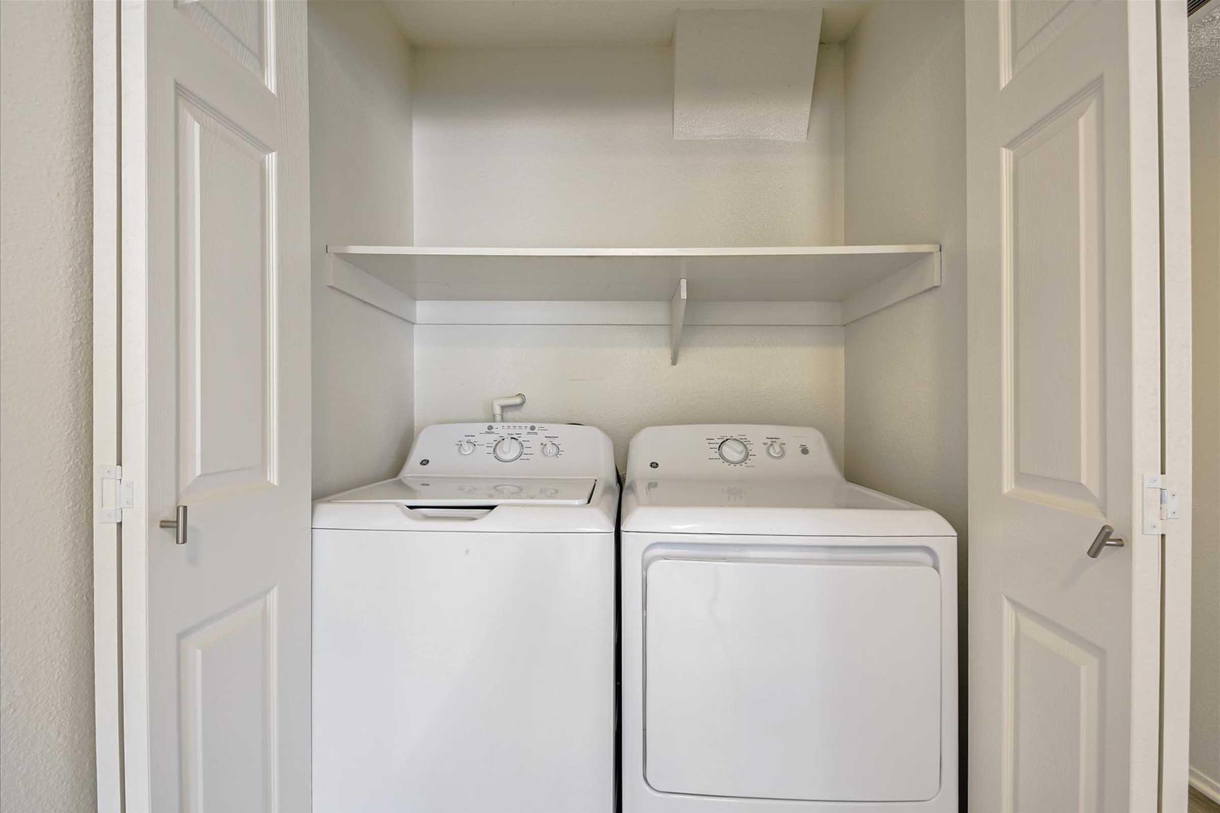 A small laundry area in a closet featuring a white washing machine and a white dryer side by side. Above the appliances, there is a shelf for storage. The closet doors are partially open, revealing the clean, light-colored interior walls.