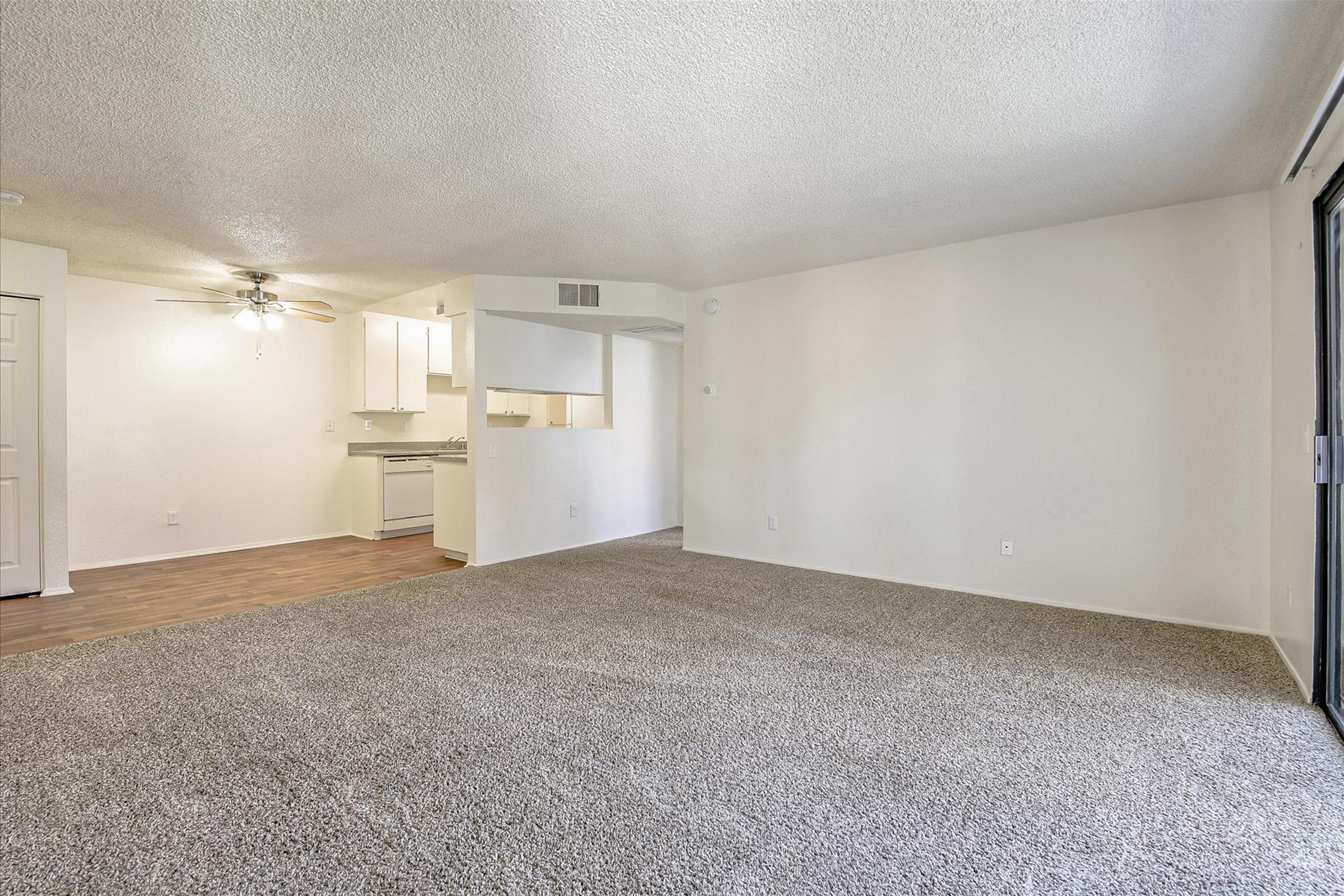 A spacious, empty living area featuring beige carpet, a ceiling fan, and an open layout. A kitchen is visible in the background with white cabinets. Large windows allow natural light, and the walls are painted in a light color, creating a bright and airy atmosphere.