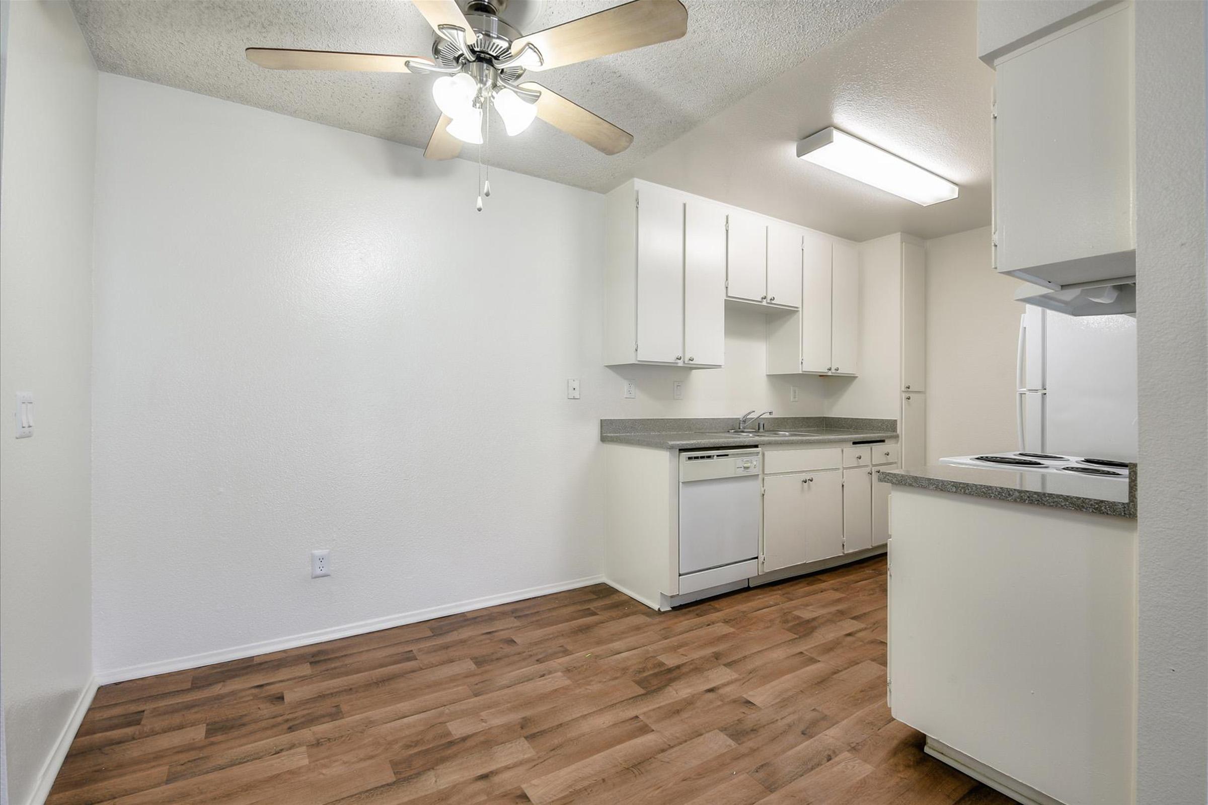 A bright, clean kitchen featuring white cabinets, a dishwasher, and a ceiling fan. The floor is made of light wooden planks, and there is a spacious layout with counter space and a refrigerator. Natural light from a window creates a welcoming atmosphere.