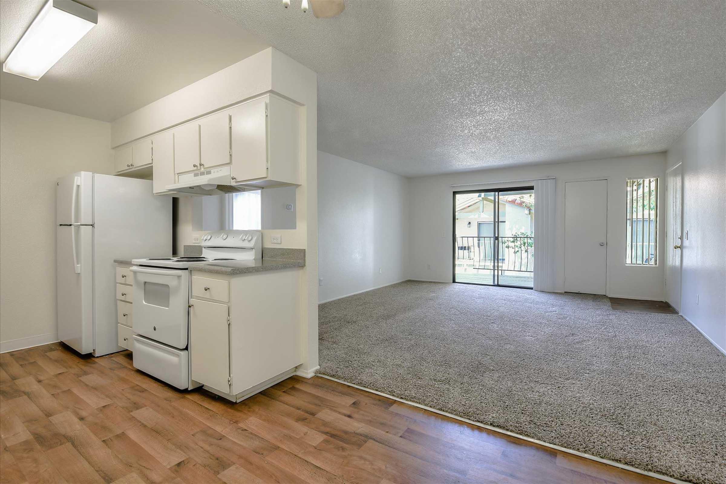 A bright, spacious living area featuring an open kitchen with white cabinets, a refrigerator, and a stove. The living room has carpeted flooring and large windows overlooking a patio. The walls are painted in neutral tones, creating a welcoming atmosphere.