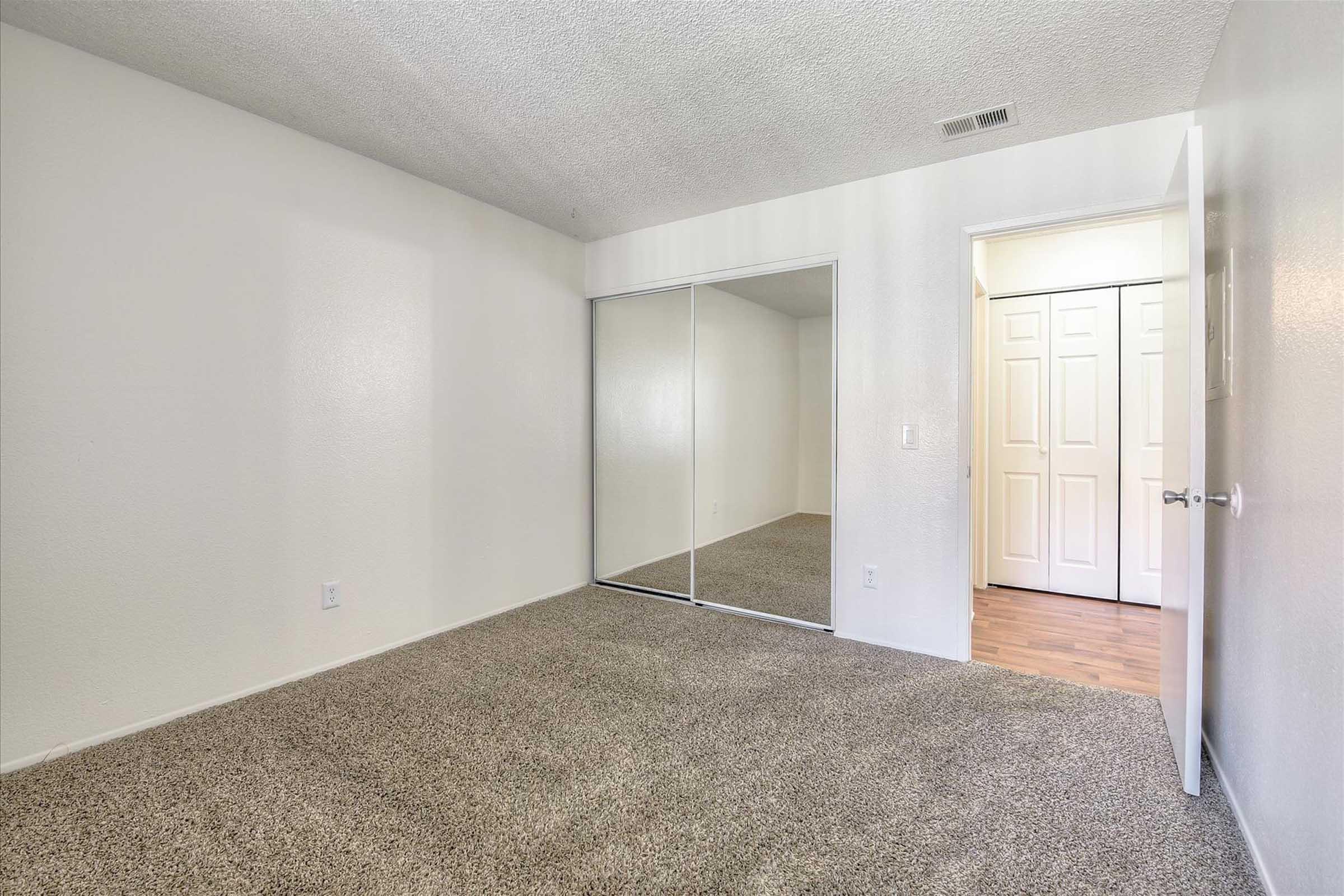 A vacant room with beige carpet, white walls, and a large mirrored closet. There is a doorway leading to another space, and the lighting appears bright and neutral, creating a clean and open atmosphere.