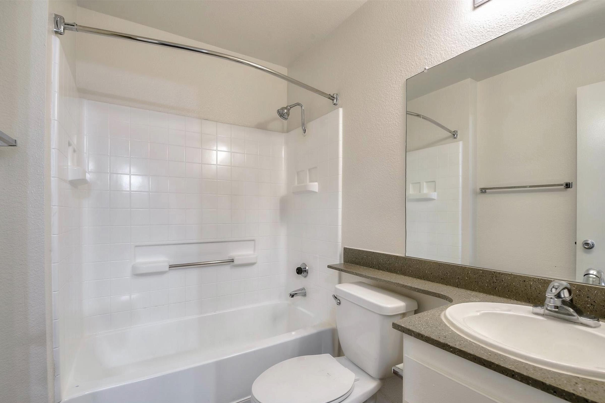A clean, well-lit bathroom featuring a bathtub with a shower, a toilet, and a sink with a granite countertop. The walls are painted white, and there is a large mirror above the sink. The overall aesthetic is modern and minimalistic.