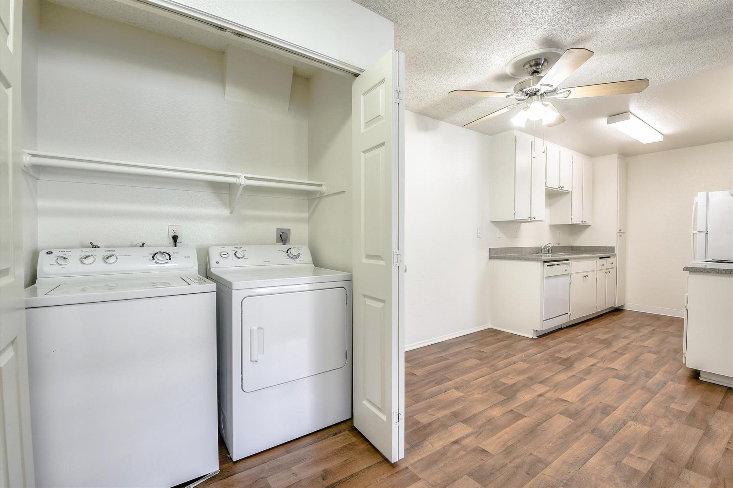 A bright laundry area featuring a washer and dryer, located next to a modern kitchen. The kitchen includes white cabinets, a countertop, and appliances like a refrigerator and dishwasher. The room has light-colored walls and wooden flooring, complemented by a ceiling fan.
