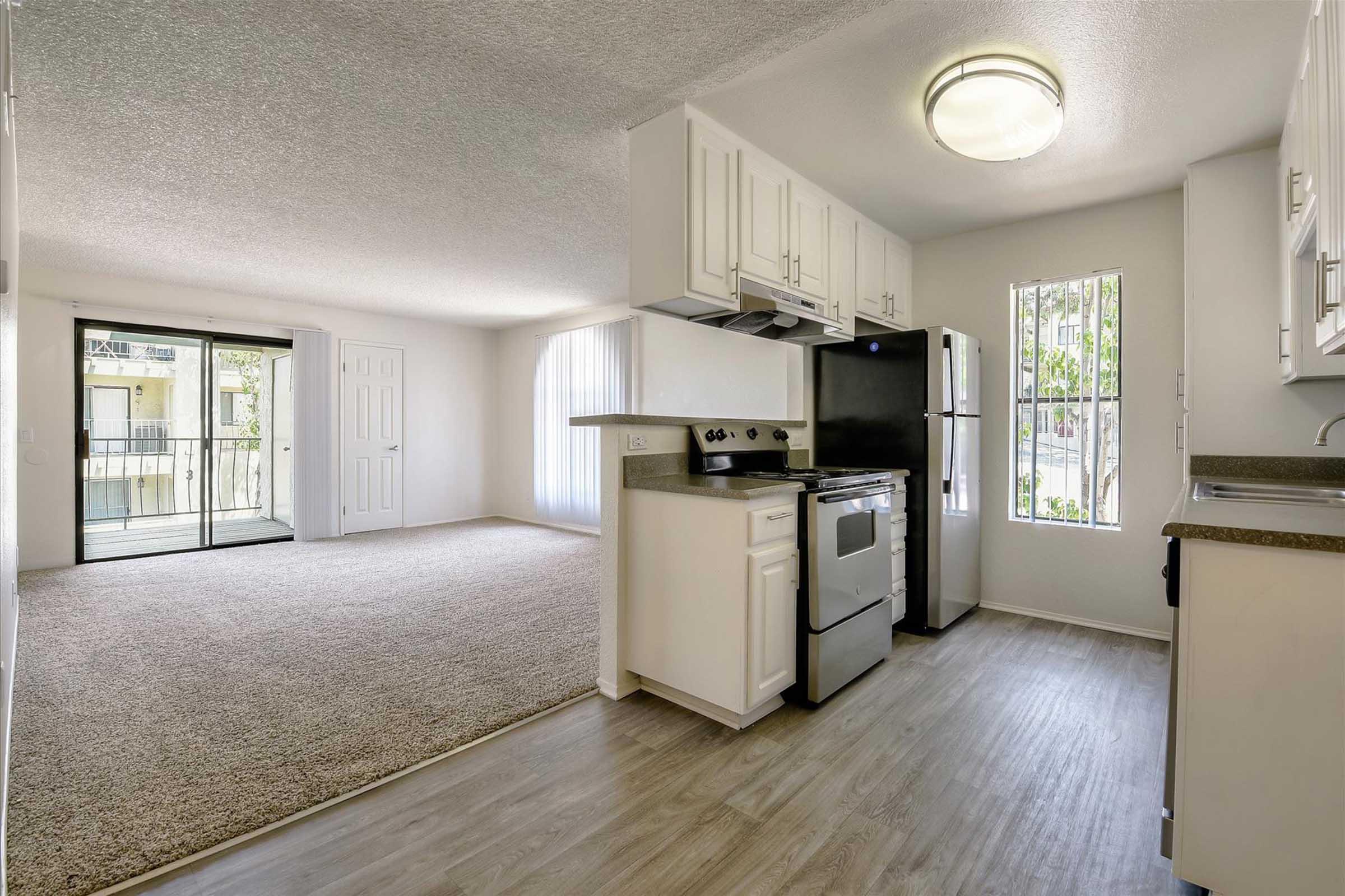 A modern kitchen with white cabinetry, a black refrigerator, and a silver stove, featuring a countertop. The space opens into a bright living area with a sliding glass door leading to a balcony. The flooring is light-colored, and there is plush carpet in the living area, creating a spacious and inviting atmosphere.