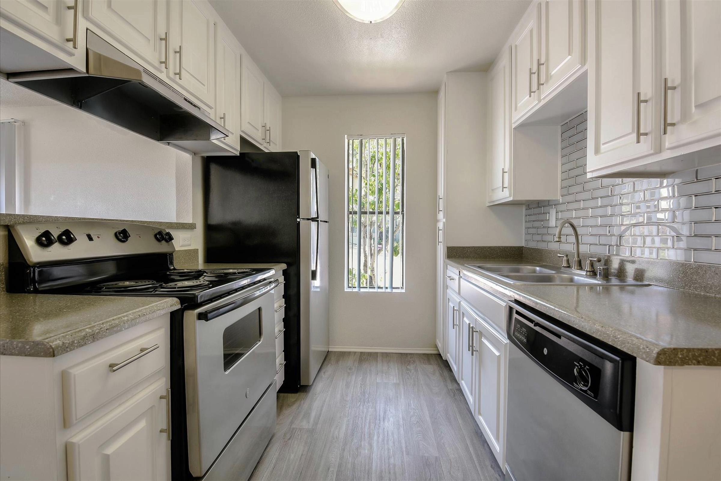 Modern kitchen featuring white cabinetry, stainless steel appliances, and granite countertops. Includes a black refrigerator, stove, and dishwasher. The design is sleek with a window providing natural light and a backsplash of white subway tiles. The floor is light-colored wood laminate.