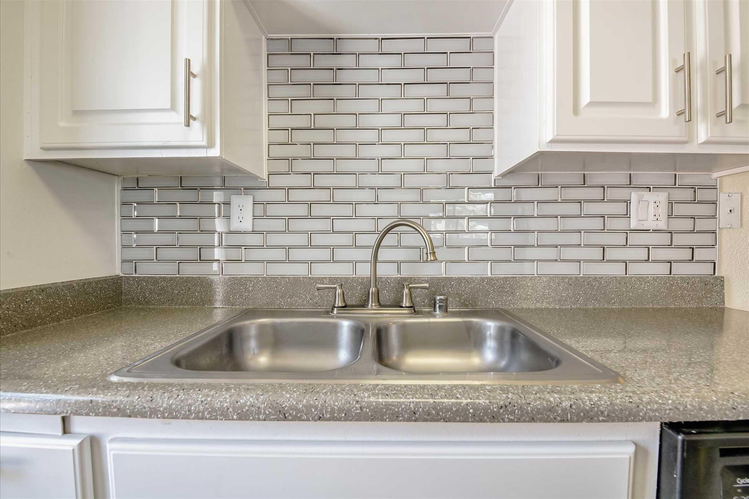 A modern kitchen sink area featuring a double stainless steel sink with a sleek faucet, set against a stylish backsplash of grey subway tiles. The countertop is a light-colored speckled design, and white cabinetry frames the space, providing a clean and contemporary look.