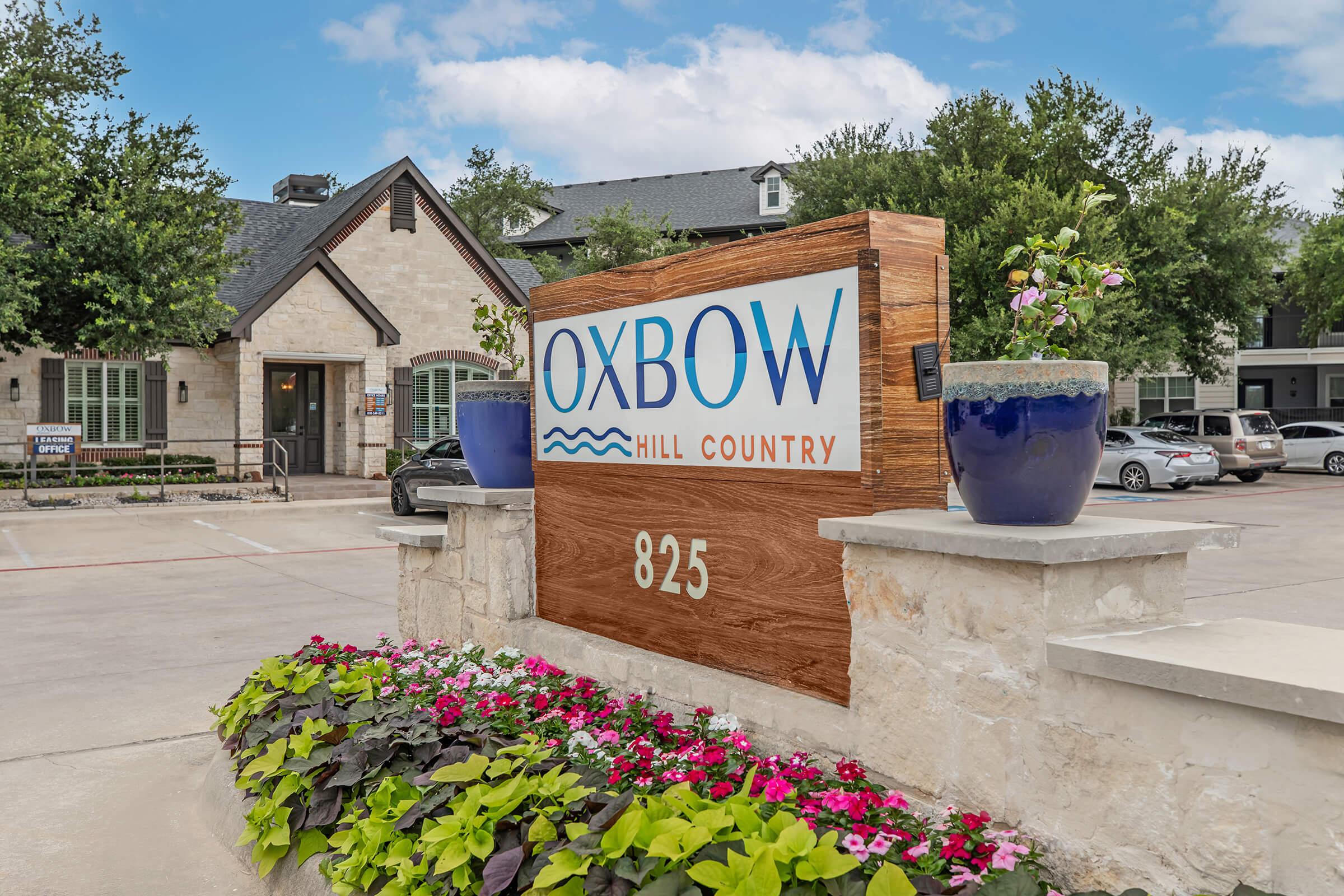 Signage for Oxbow Hill Country at 825, featuring a modern design with blue text against a wooden background. Surrounding the sign are vibrant planters filled with colorful flowers, and the building's exterior is visible in the background under a partly cloudy sky.
