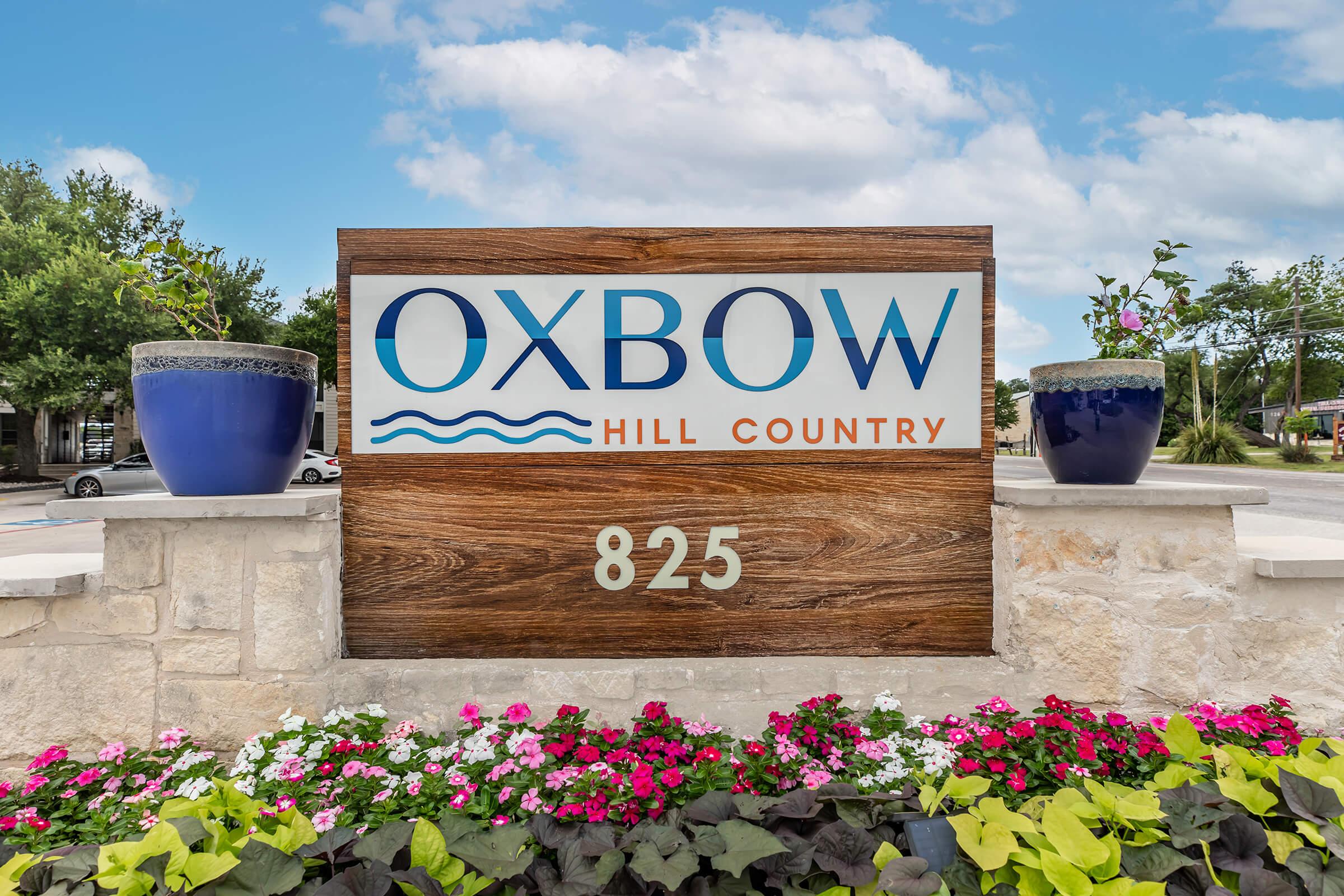 A wooden sign displaying "Oxbow Hill Country" with the number "825" beneath it. The sign is surrounded by decorative planters with blue pots and vibrant flowers, set against a backdrop of a clear sky with scattered clouds.