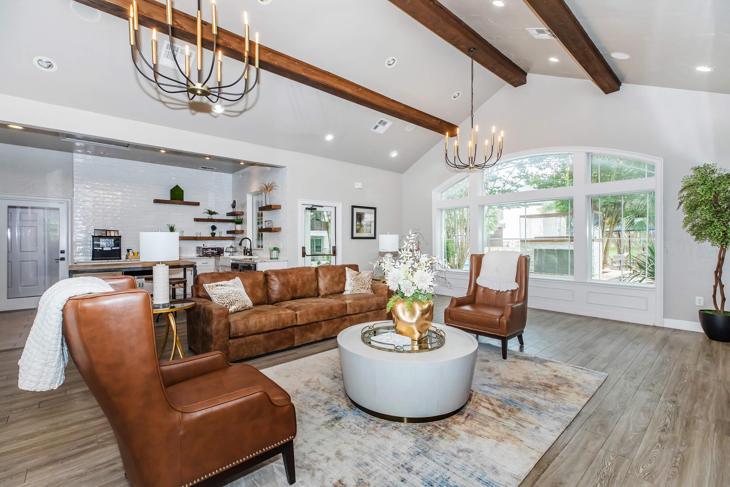 A stylish living room featuring a brown leather sofa and two armchairs, a round coffee table with a decorative vase, and large windows allowing natural light. Wood beam ceilings, neutral-colored walls, and decorative shelving add to the modern design, creating a cozy and inviting atmosphere.