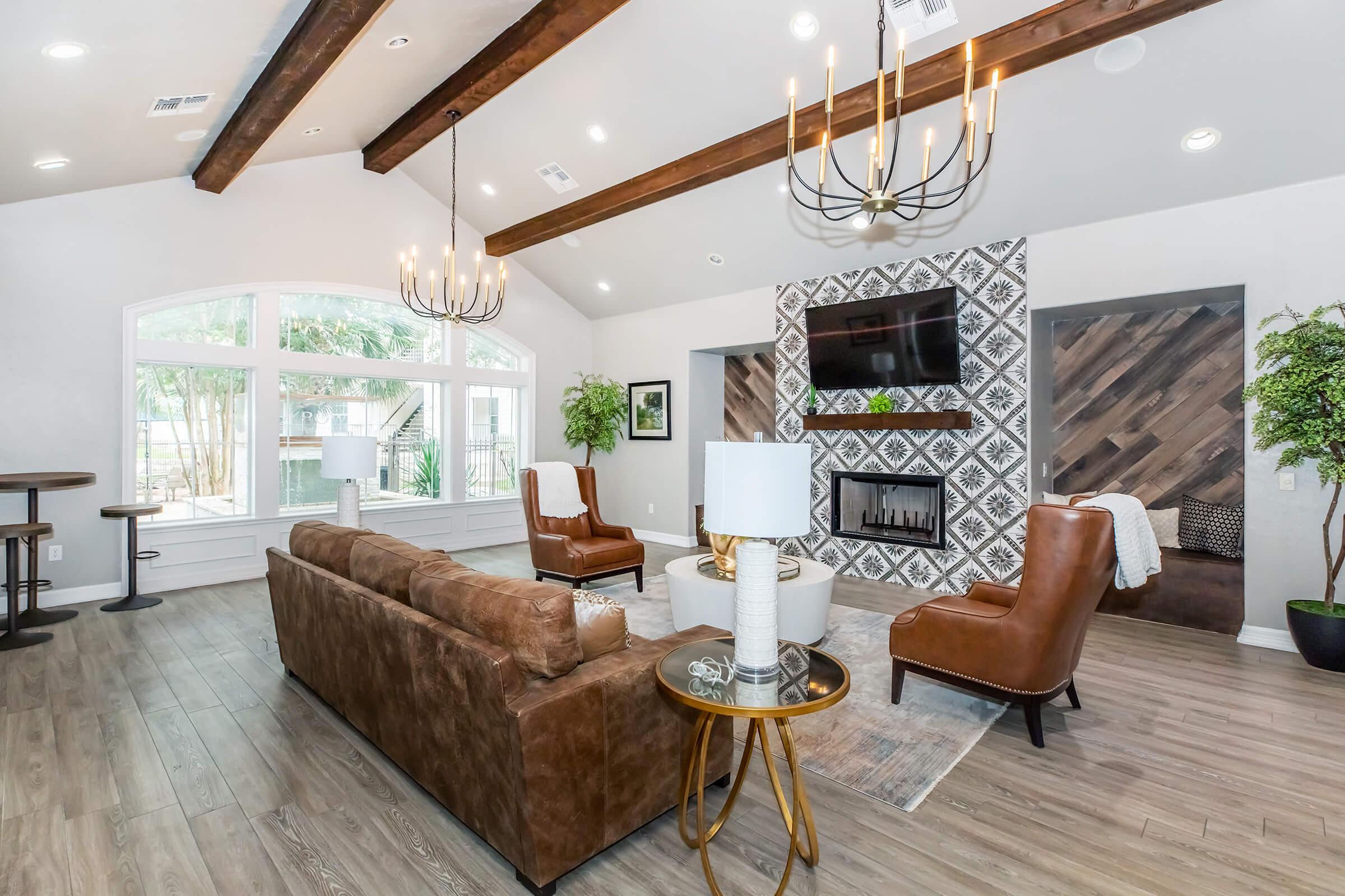 A modern living room featuring brown leather chairs and a sectional sofa, a round coffee table, and a decorative lamp. The space has high ceilings with exposed wooden beams, a large wall-mounted TV above a stylish fireplace, and large windows providing natural light, enhanced by greenery in the decor.