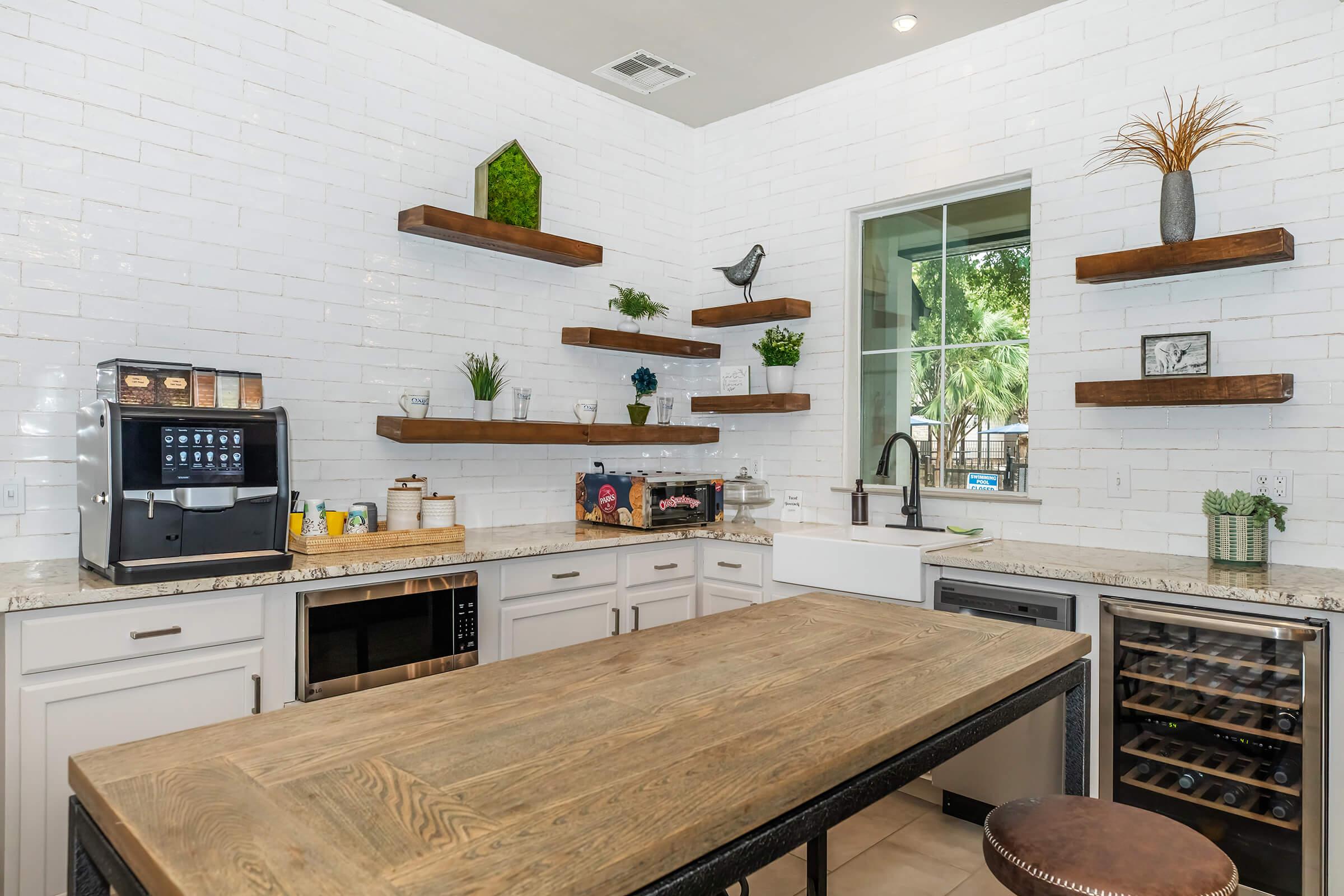Modern kitchen with white brick backsplash, featuring wooden shelves with decorative plants and a bird figurine. The kitchen includes a coffee machine, microwave, and wine cooler. A large wooden island with a metal frame is central, complemented by natural light from a nearby window.