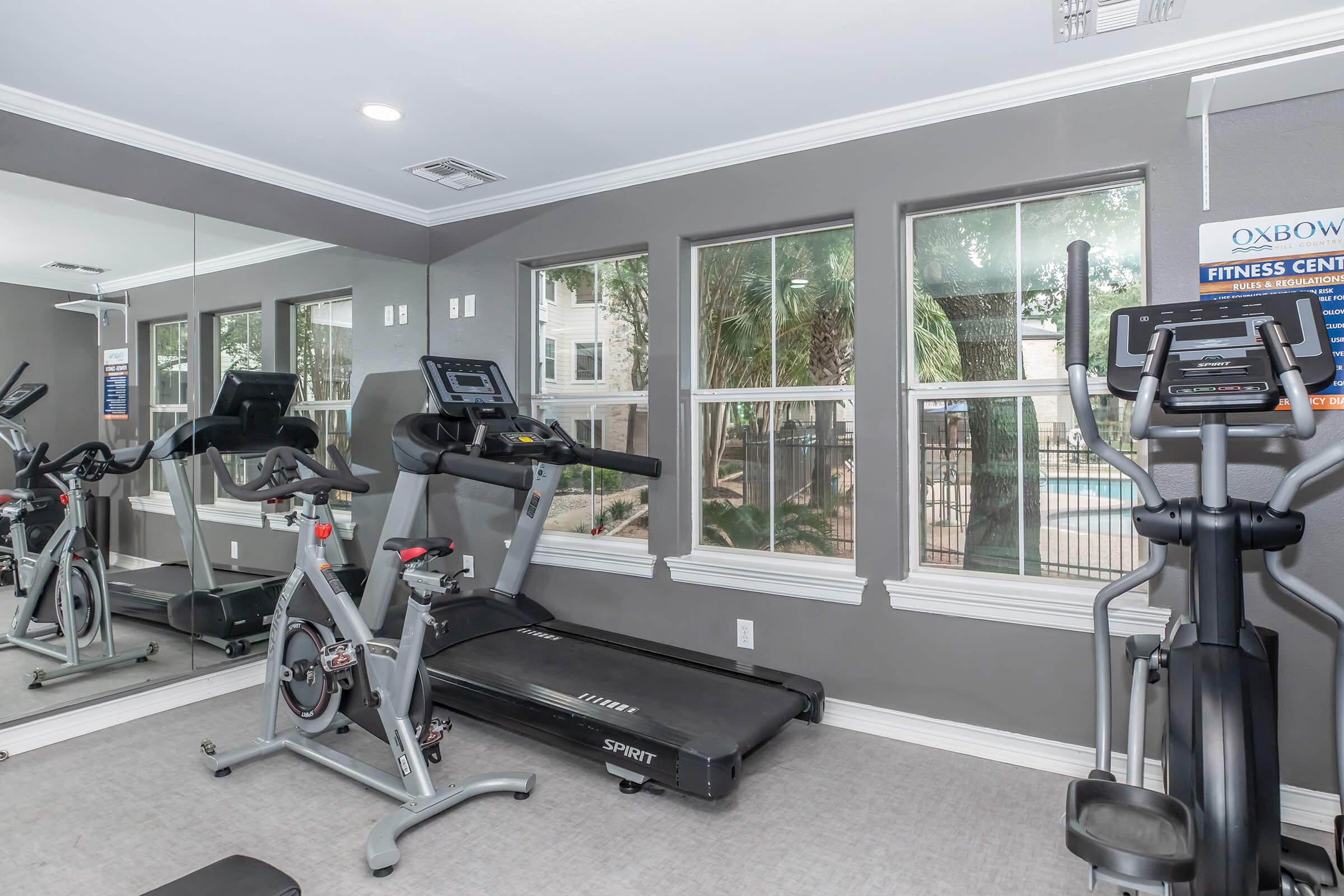 A well-lit fitness center with large windows showing a view of the outdoors. The room features a treadmill and an elliptical machine, with a mirror on one wall. The decor is modern with neutral colors, creating an inviting workout space.