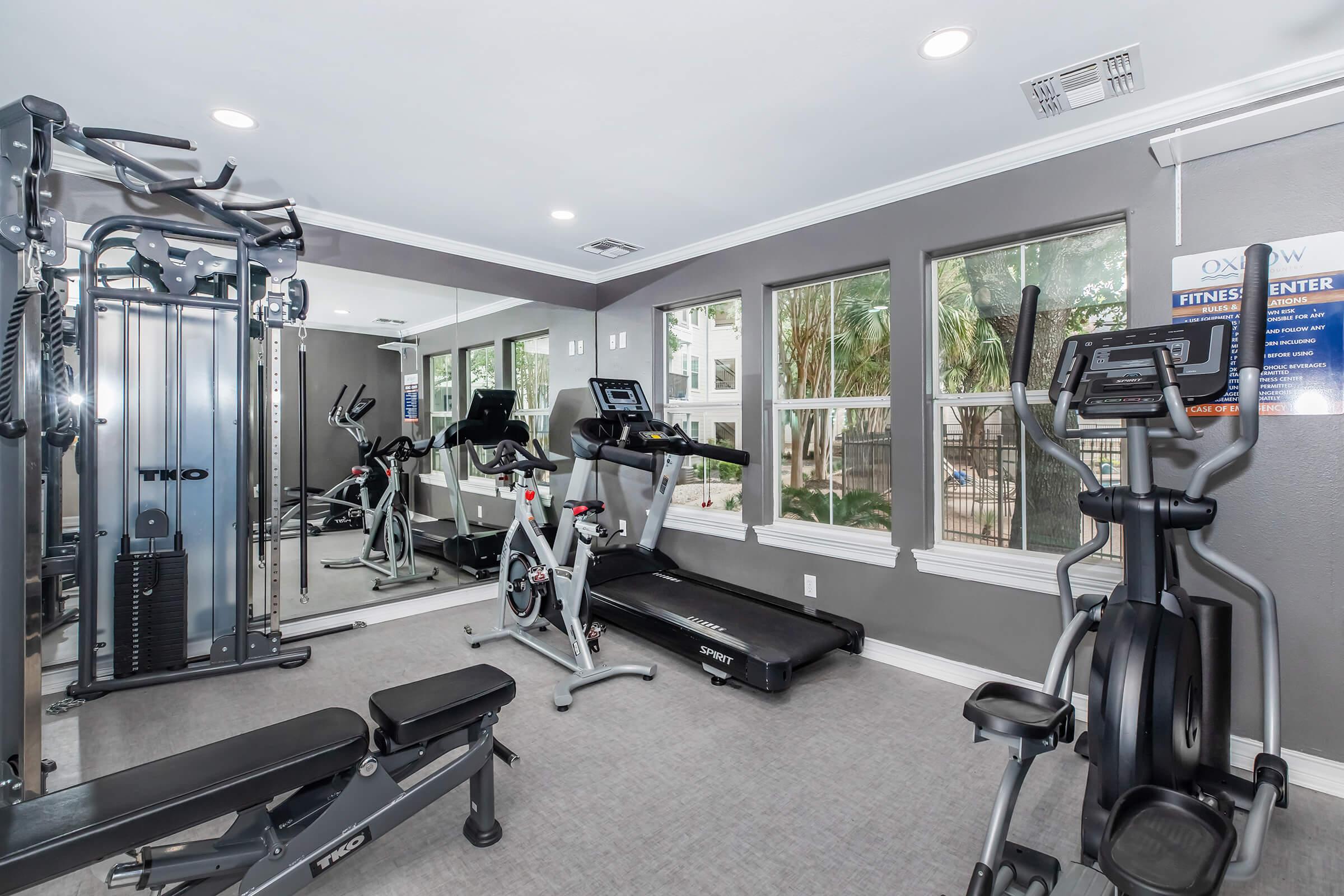 A well-lit fitness center featuring various exercise equipment, including a treadmill, stationary bike, weight bench, and a multi-gym machine. Large windows provide natural light and a view of greenery outside. Mirrors on the walls enhance the sense of space.