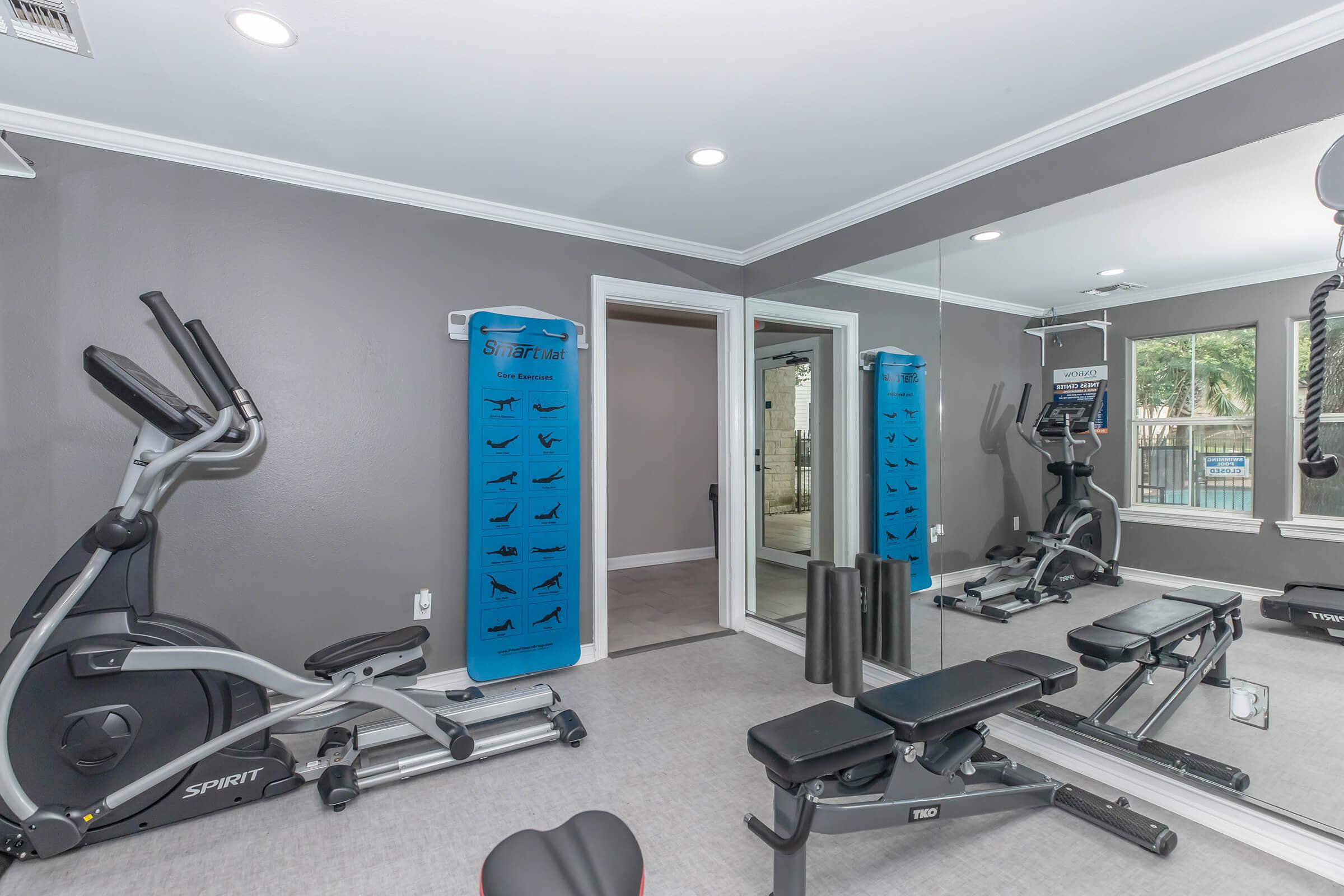 A modern home gym featuring an elliptical machine, weight bench, and mirrored walls with a poster displaying various workout exercises. The color scheme is neutral with gray walls and bright lighting, creating an inviting workout space. A door leads to another area, enhancing accessibility.