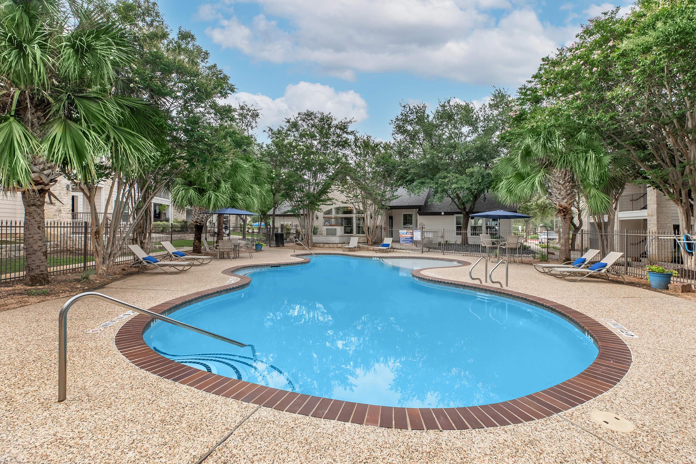 A swimming pool surrounded by palm trees and lounge chairs, set in a landscaped area. The pool features a gently curved shape with a shallow entry. In the background, there are residential buildings and umbrellas offering shade. The sky is partly cloudy, adding to the serene atmosphere.