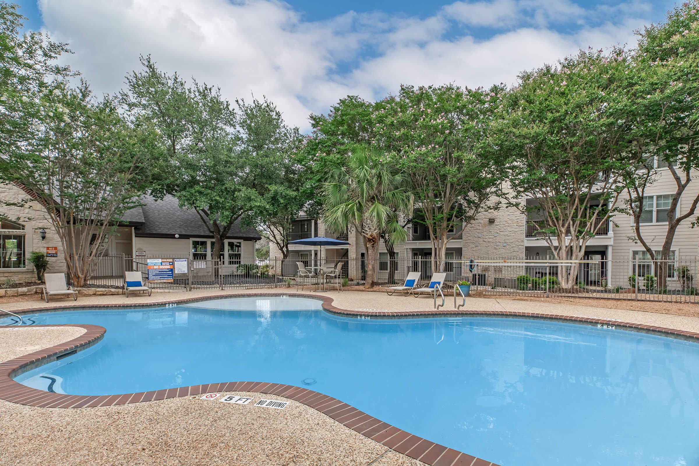 A serene outdoor swimming pool surrounded by lush greenery and a well-maintained patio area. Loungers are placed around the pool, and there are trees providing shade. The sky is partly cloudy, creating a pleasant atmosphere for relaxation. The pool features a gentle curve and clear blue water.