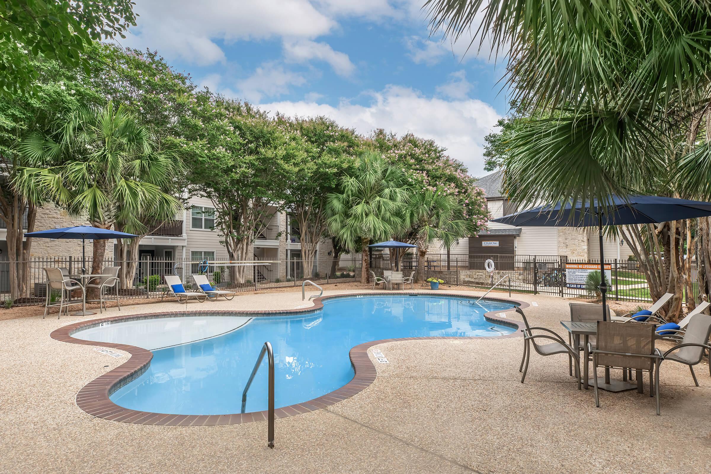 A swimming pool surrounded by palm trees and lounge chairs, with blue umbrellas providing shade. The area features a textured surface and is enclosed by a wooden fence, with a few residential buildings visible in the background beneath a partly cloudy sky.