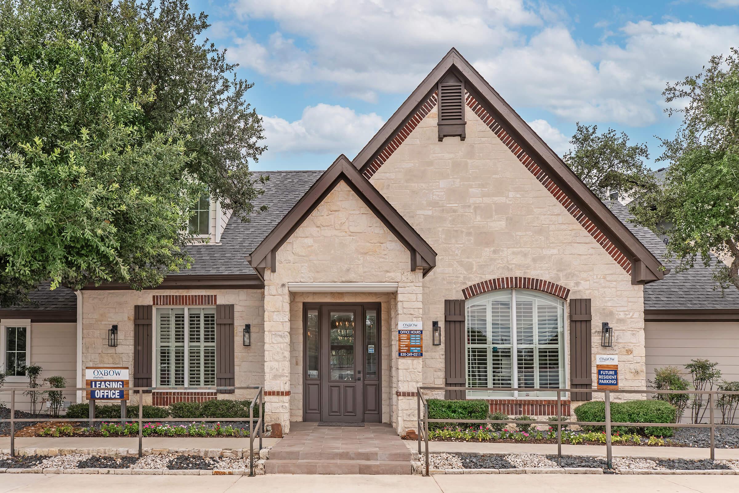A charming, single-story building made of light-colored stone and brick accents, featuring a peaked roof and large front windows. The entrance includes a welcoming door and adjacent signs indicating a leasing office. Well-maintained landscaping surrounds the property with trees and decorative stones.