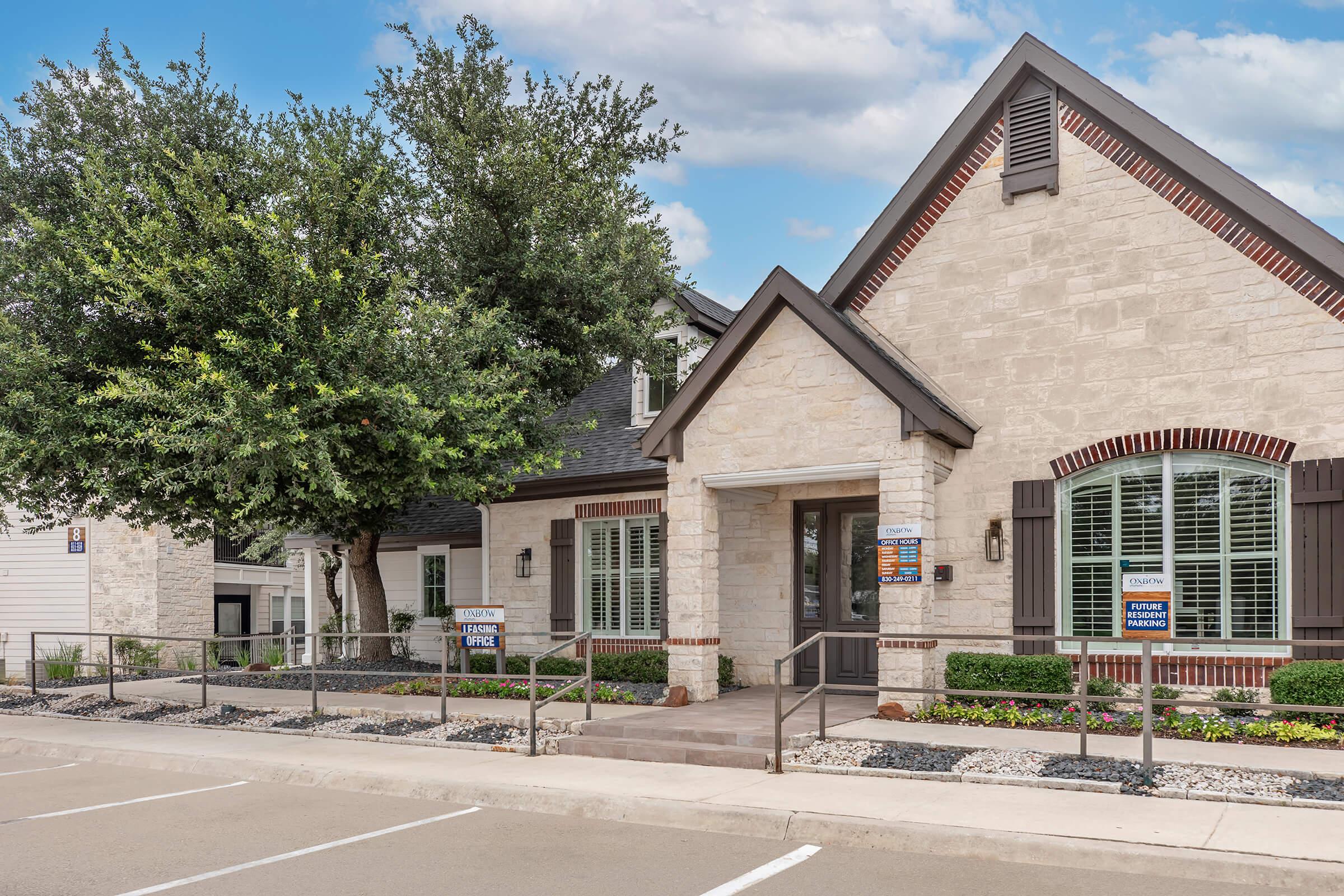 A stone building with a pitched roof featuring decorative brick accents. The entrance is framed by large windows and flanked by well-maintained landscaping, including a large tree. Signs are visible in the windows, indicating it's a property office. The scene is set against a partly cloudy sky.