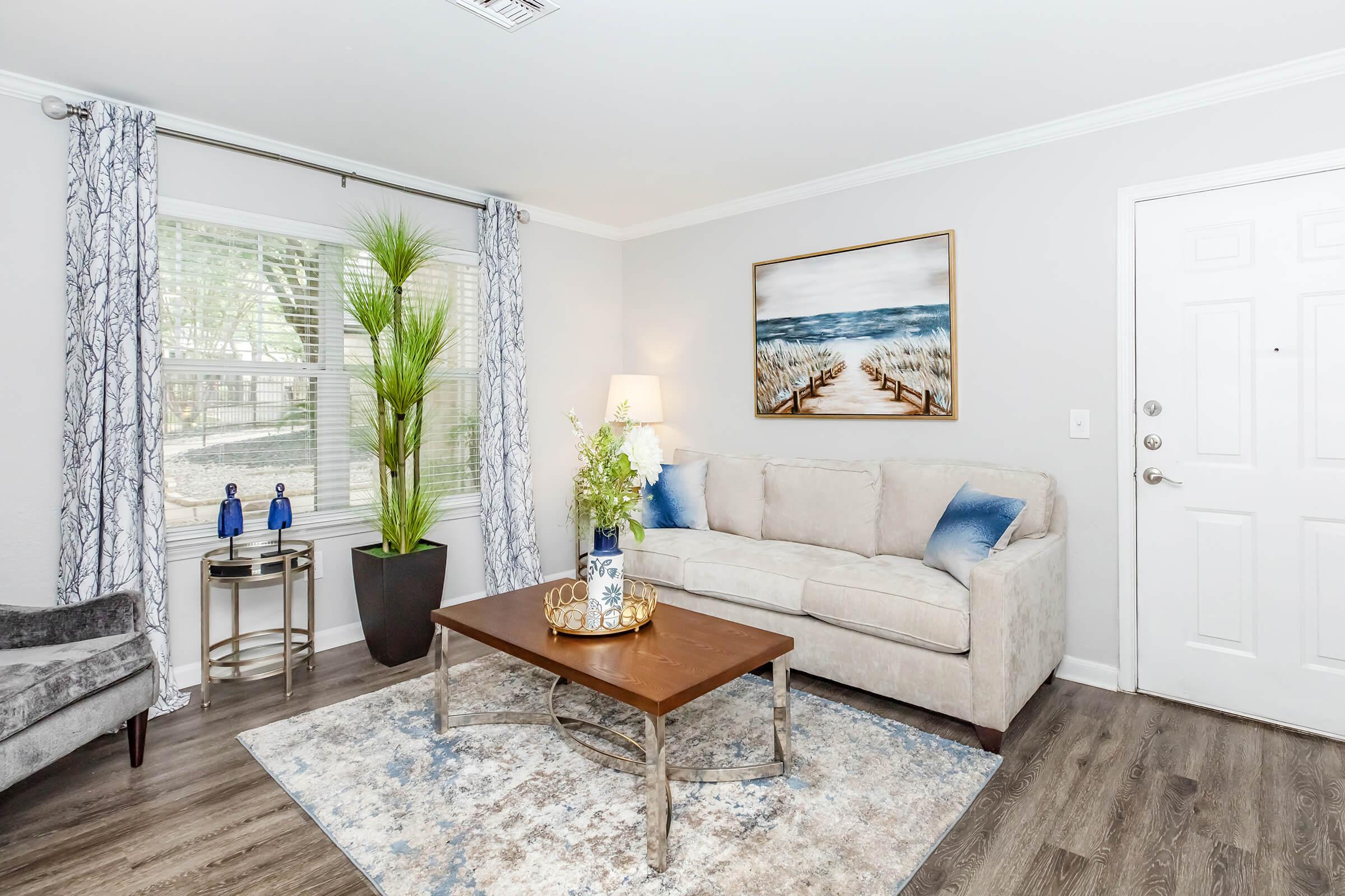 A cozy living room featuring a light gray sofa with blue accent pillows, a wooden coffee table with decorative items, and a large potted plant. The room has soft, natural light coming through the windows, with blue and white curtains and an artwork depicting a coastal scene on the wall.