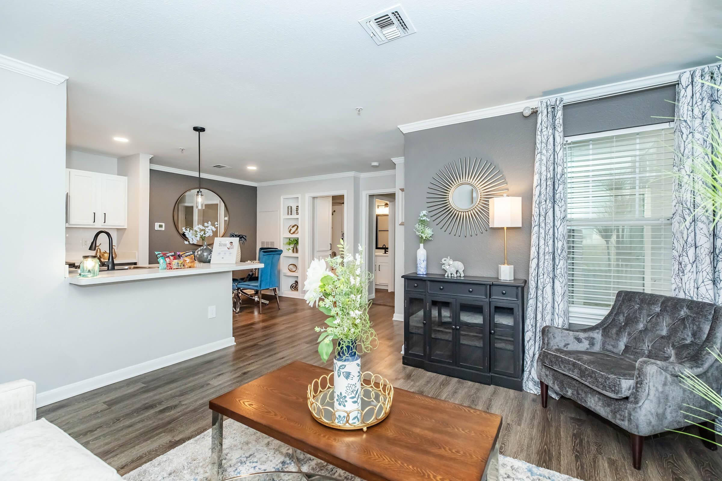 A modern living room featuring a stylish coffee table with a decorative vase, a comfortable gray armchair, and a black console table. In the background, there is a kitchen area with white cabinets and a bar seating area. Large windows are dressed with patterned curtains, adding brightness to the space.