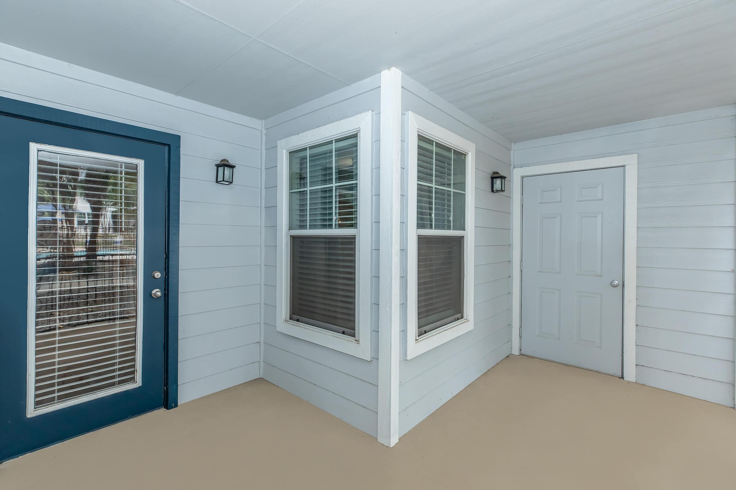 A corner view of a light blue exterior with two windows and a blue door on the left. The walls are made of paneling, and there is a light fixture next to the windows. A plain white door is visible on the right side, completed with a beige floor.