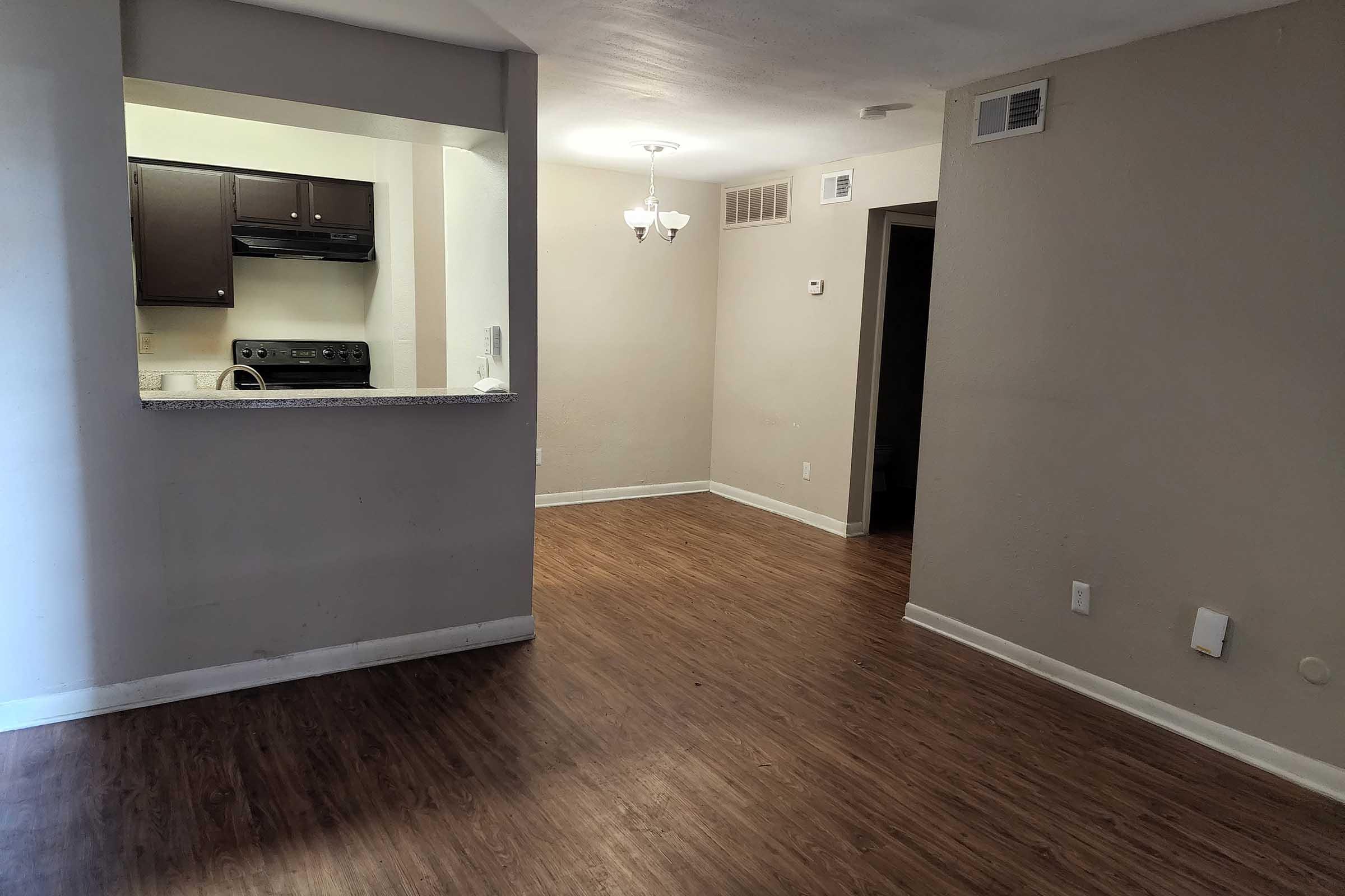 A spacious living area with hardwood-style flooring, a small dining area lit by a chandelier, and an open kitchen visible in the background. The walls are painted in a neutral tone, and there are doorways leading to other rooms. The kitchen features dark cabinetry and appliances.