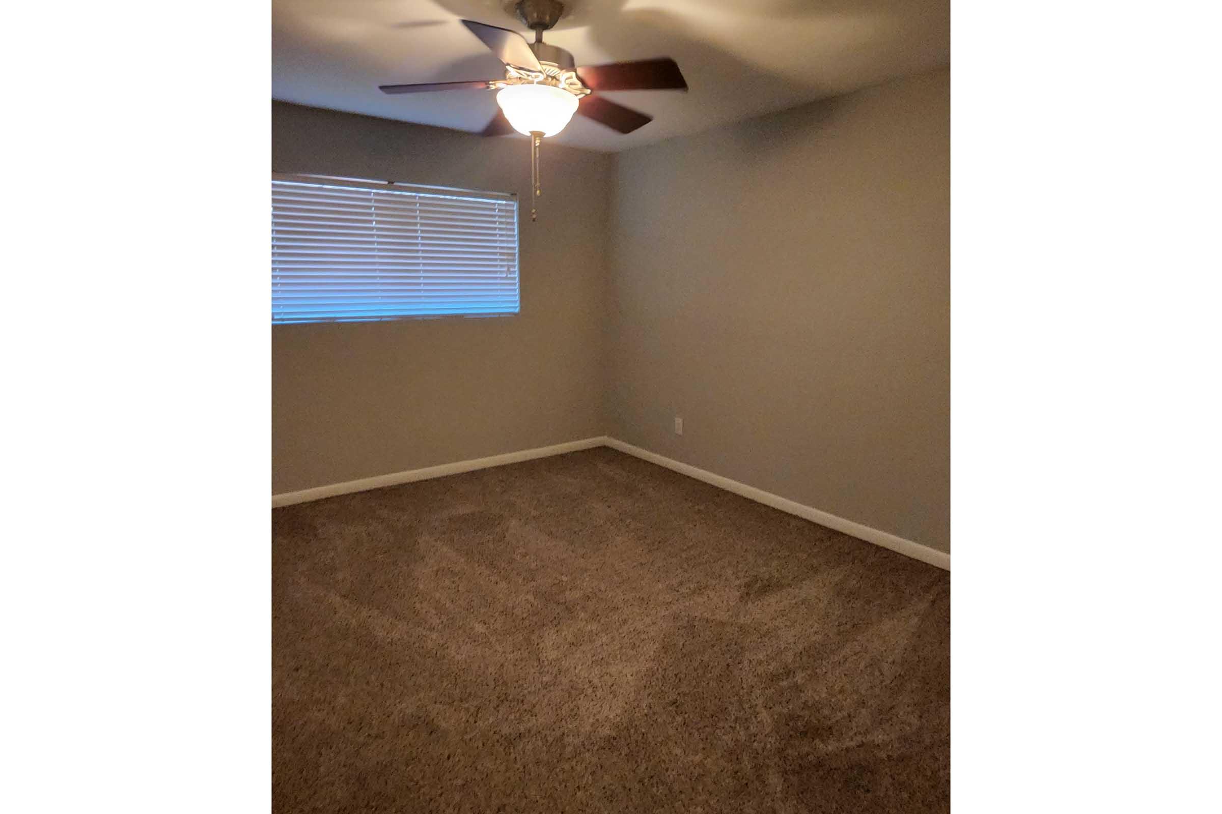 A simple, empty room with light gray walls and a ceiling fan. The floor is covered with carpet, and there is a window with closed blinds allowing limited light. The overall atmosphere is calm and inviting, ideal for various uses.