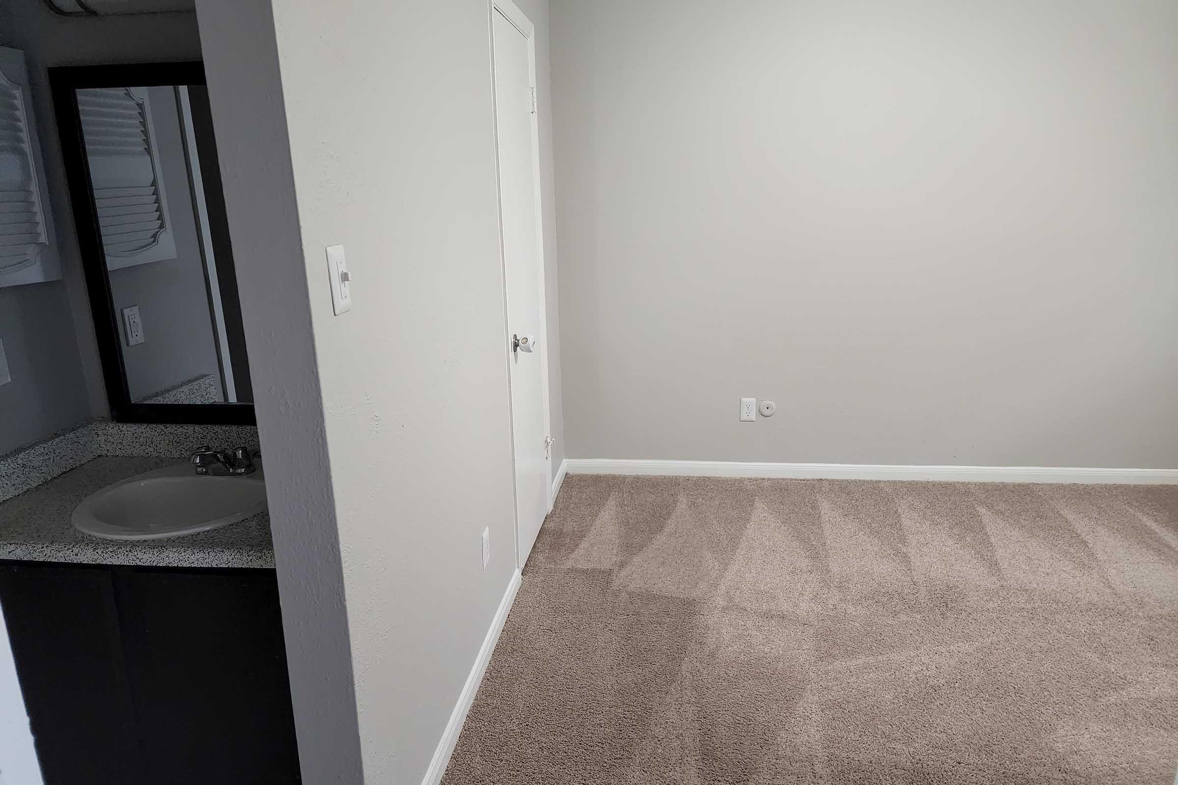 A view of a small, empty room with light gray walls and a carpeted floor. There is a doorway leading to another space and a bathroom area visible with a sink and a mirror. The lighting is bright, creating a spacious feel in the room.