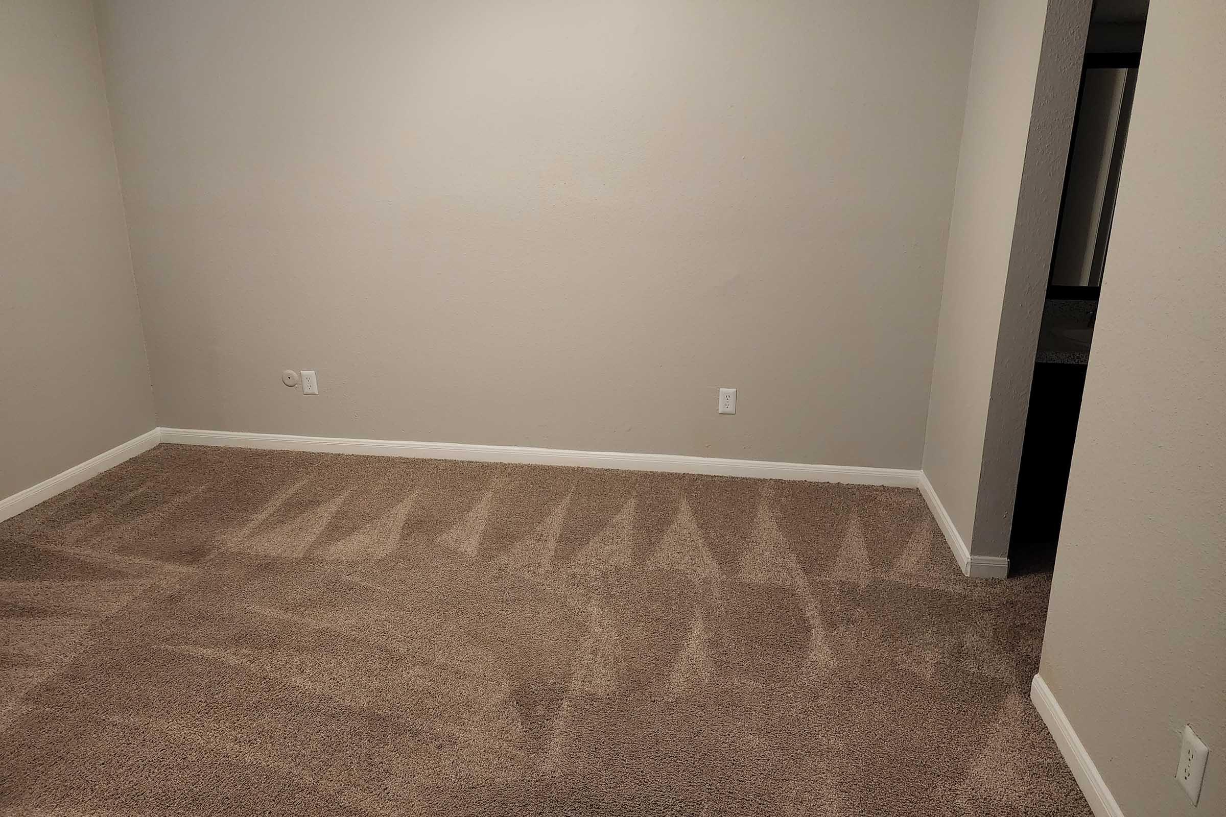 Empty room with light beige walls and carpeted flooring. The carpet appears to have marks from furniture. There is a doorway visible leading to another space, with natural light illuminating the area. No furniture or decorations are present.