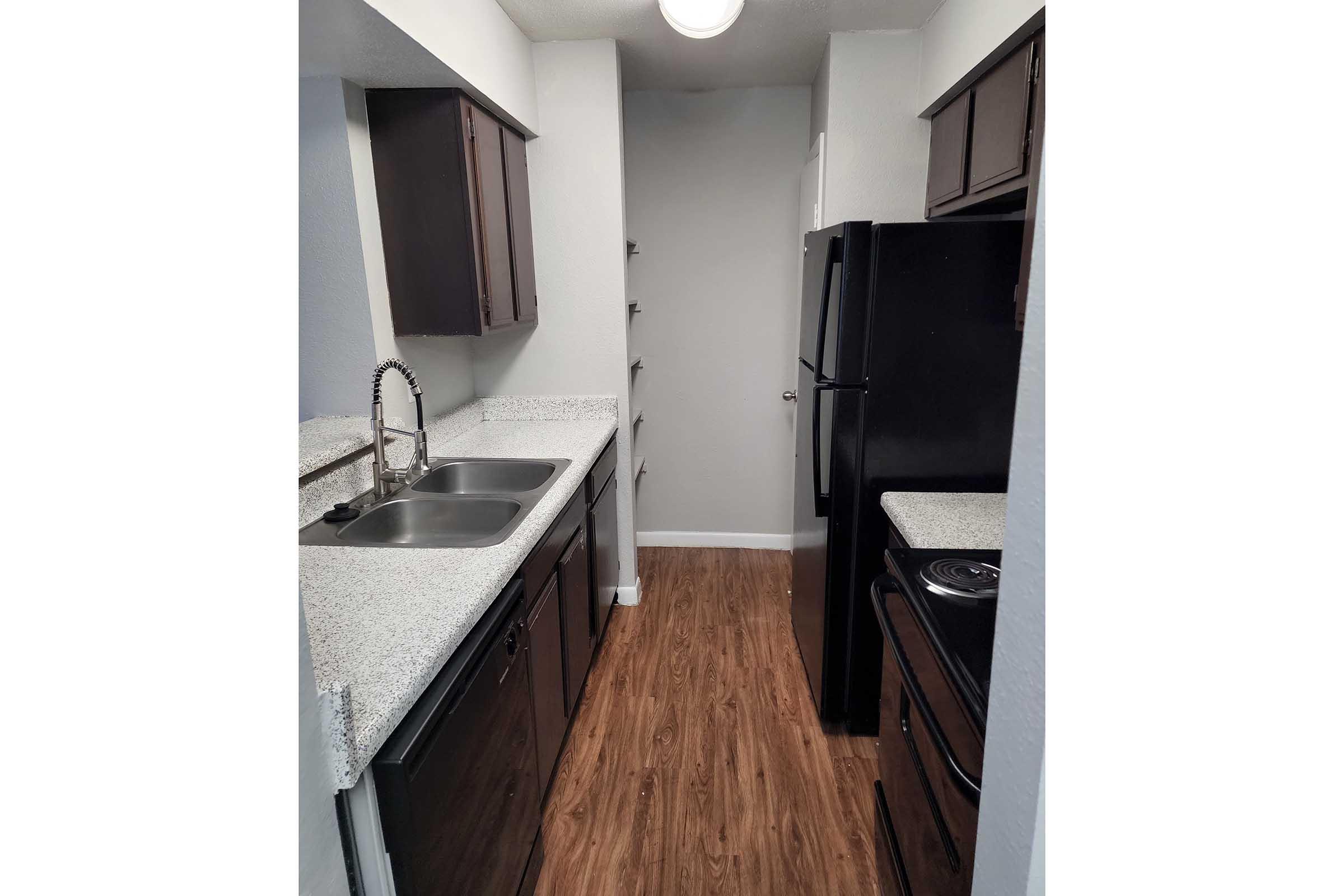 A modern kitchen featuring sleek dark cabinets, a double sink, and a countertop with a speckled design. The appliances include a black refrigerator and stove. There’s a narrow corridor leading to a door at the back, with additional storage shelves on the wall. The floor is made of wood laminate.