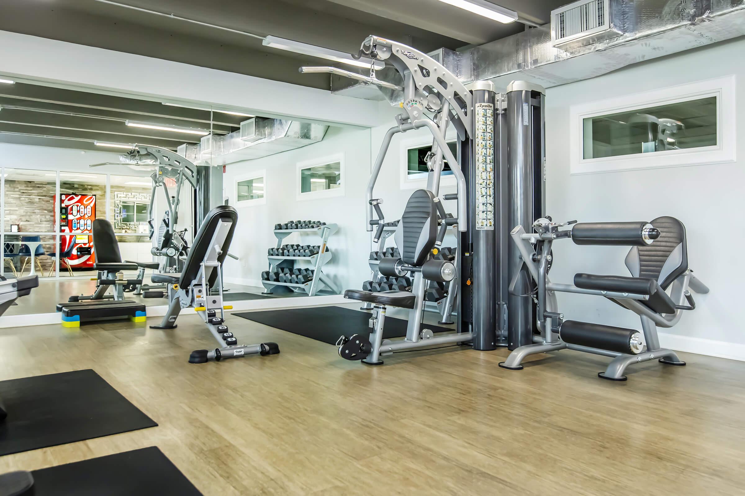 A modern gym interior with various exercise equipment, including weight machines and free weights. The space features large mirrors, wooden flooring, and a clean, well-lit environment, creating an inviting atmosphere for workouts.