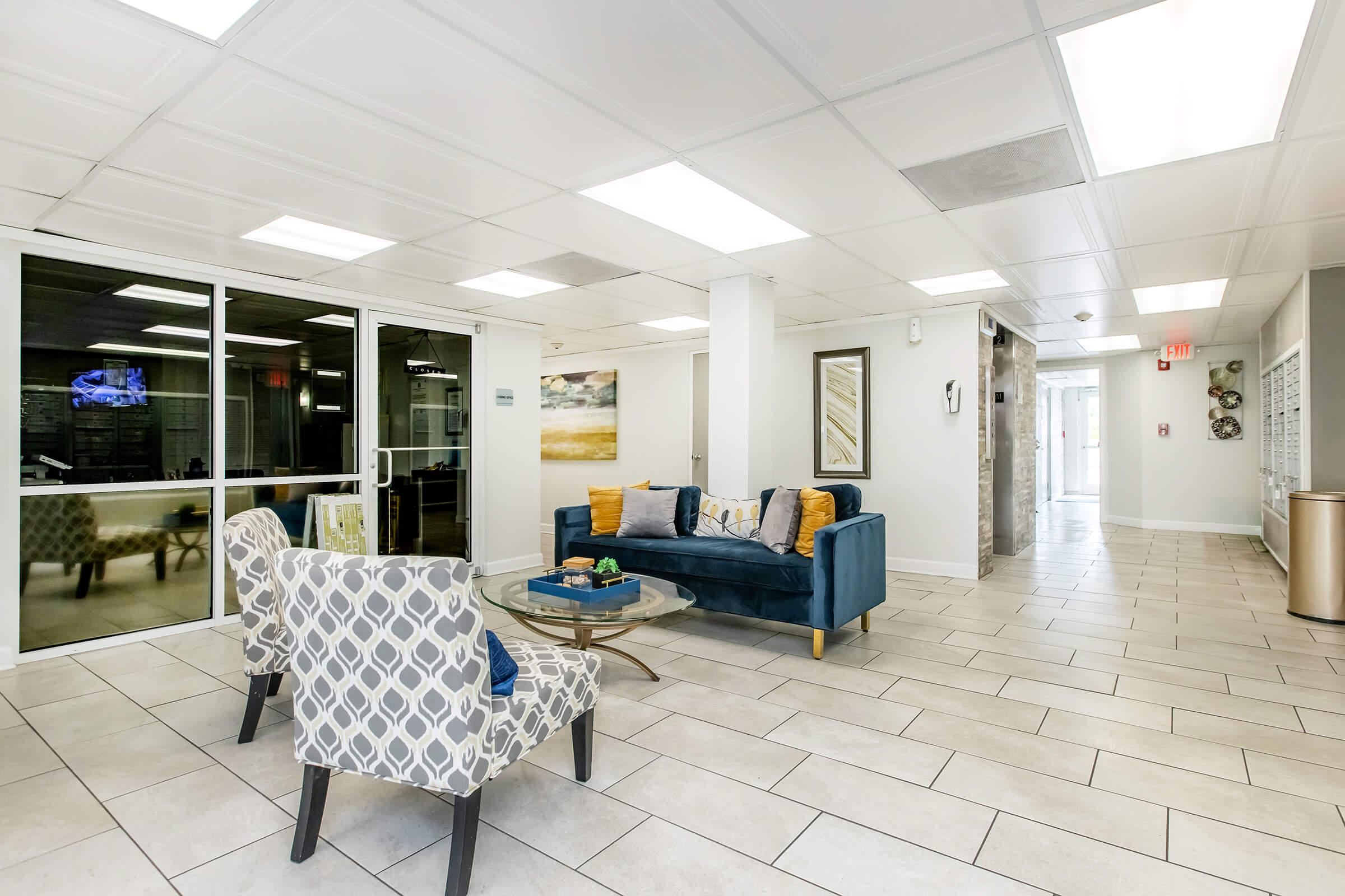 A modern lobby area featuring a blue sofa with decorative pillows, two patterned chairs, and a glass coffee table. The space has tile flooring, bright overhead lighting, and large windows allowing natural light. There are artworks on the walls and a television visible in the background.