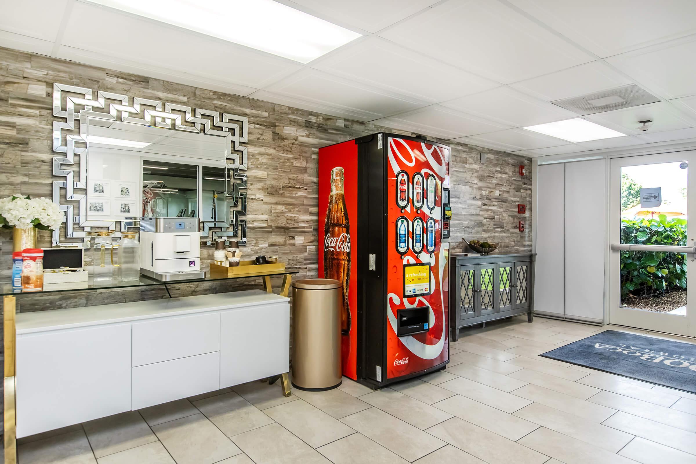 A modern lobby area featuring a Coca-Cola vending machine on one side, a stylish white cabinet with a trash bin nearby, and a large mirror on the wall. The floor is tiled, and there are decorative plants and furnishings that add to the welcoming atmosphere. Natural light streams in from a nearby window.