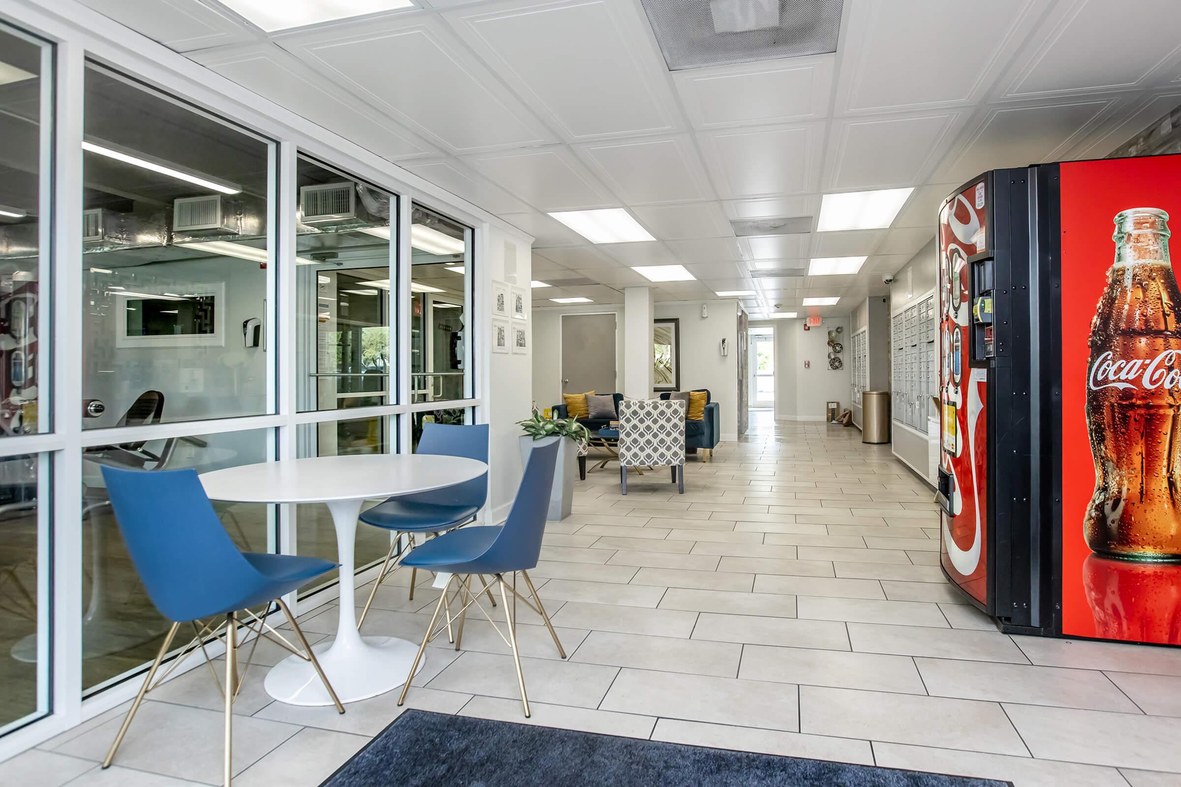 A modern interior space featuring a round white table with two blue chairs, a Coca-Cola vending machine on the right, and a bright hallway with glass partitions leading to other areas. The tiled floor adds to the contemporary design, creating a welcoming atmosphere.