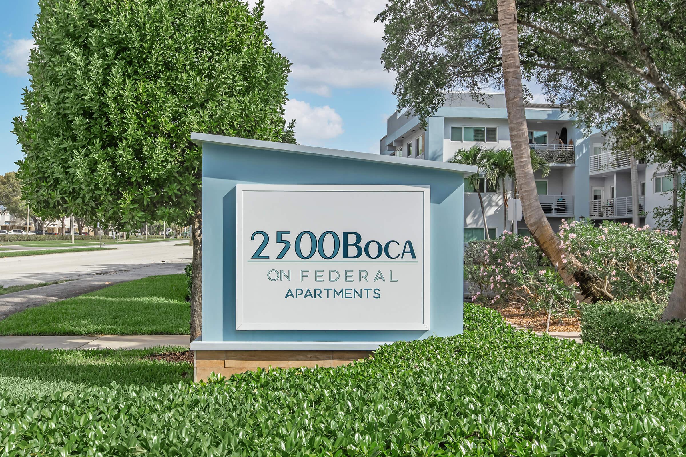 Sign for "2500 Boca on Federal Apartments" surrounded by well-maintained landscaping, featuring green bushes and trees. The background shows a road and nearby apartment buildings, indicating a residential area.