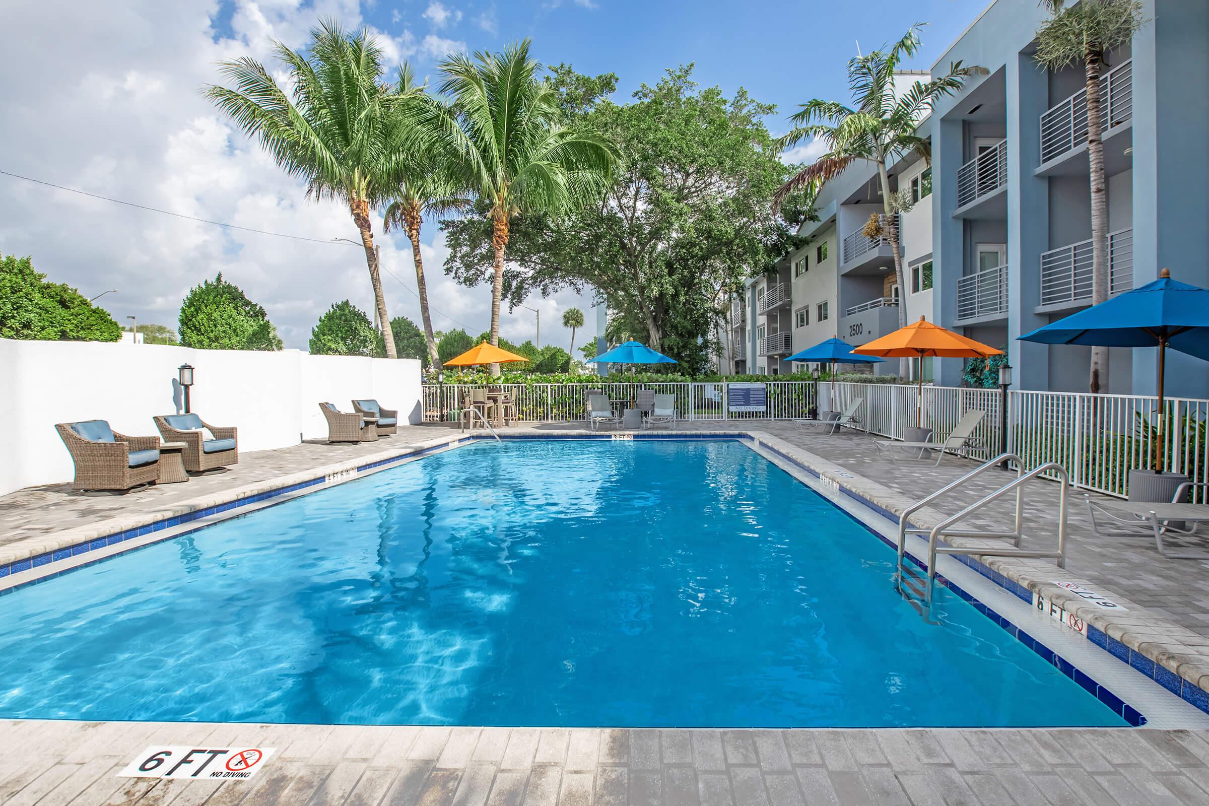 A clear blue swimming pool surrounded by lounge chairs and umbrellas in a tropical setting. Palm trees and a white wall are visible in the background, along with a multi-story building. Signs indicate a six-foot depth at one end of the pool. The scene is bright and inviting, perfect for relaxation.
