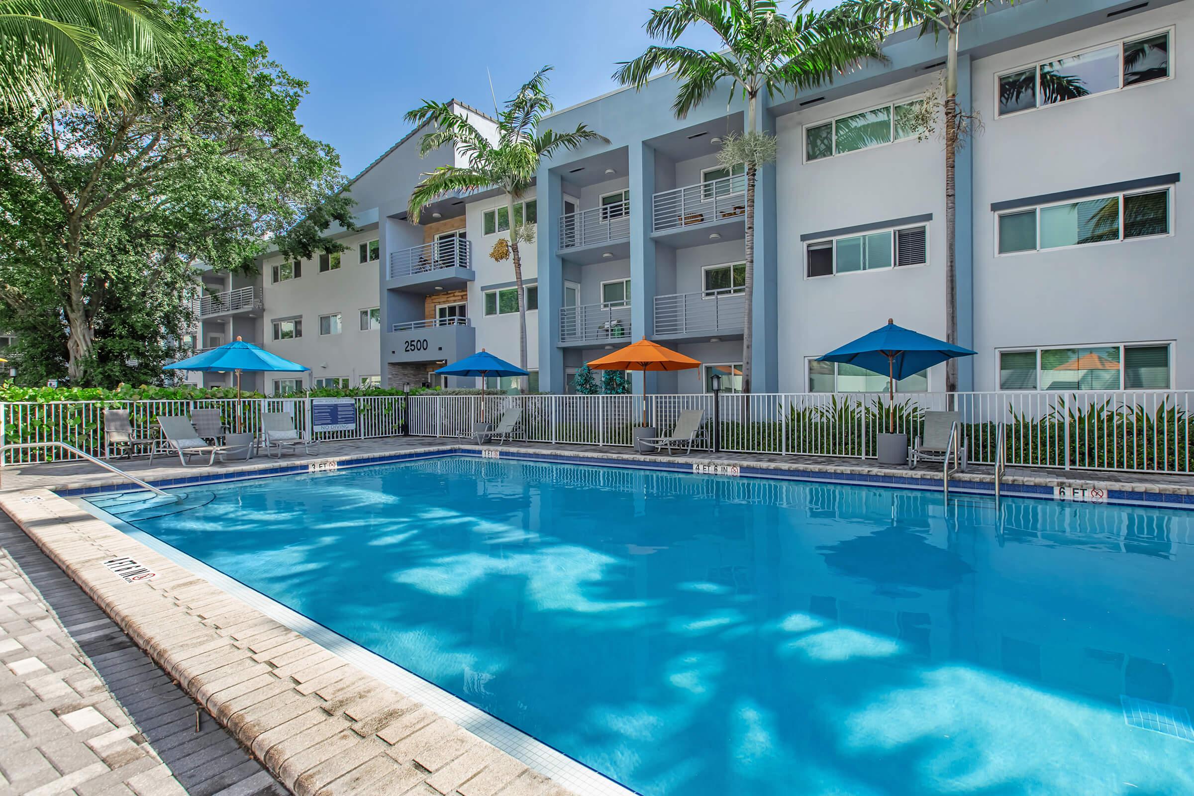 A sunny outdoor pool area surrounded by palm trees, featuring a clear blue pool with lounge chairs, and colorful umbrellas in orange and blue. Adjacent to the pool are modern apartment buildings with balconies and large windows, creating a relaxing atmosphere.