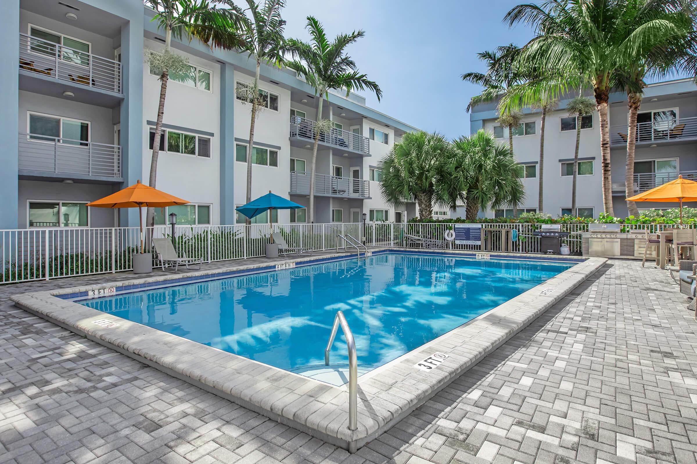 A serene outdoor swimming pool surrounded by palm trees and apartment buildings. The pool features a shallow area with a ladder, and there are several orange umbrellas providing shade. The area is paved with light-colored bricks, creating a relaxing atmosphere for residents.