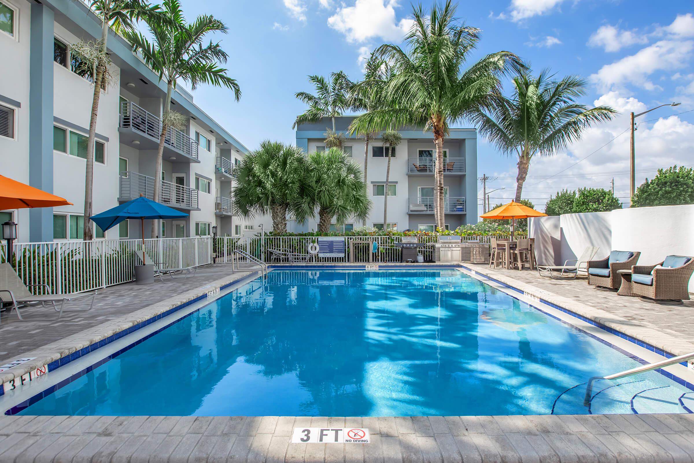 A tranquil pool area surrounded by palm trees and lounge chairs. The pool features a clear blue water surface, with umbrellas providing shade. Adjacent buildings are visible in the background, and a "3 FT" depth marker is positioned at the pool's edge. Bright, sunny weather enhances the inviting atmosphere.