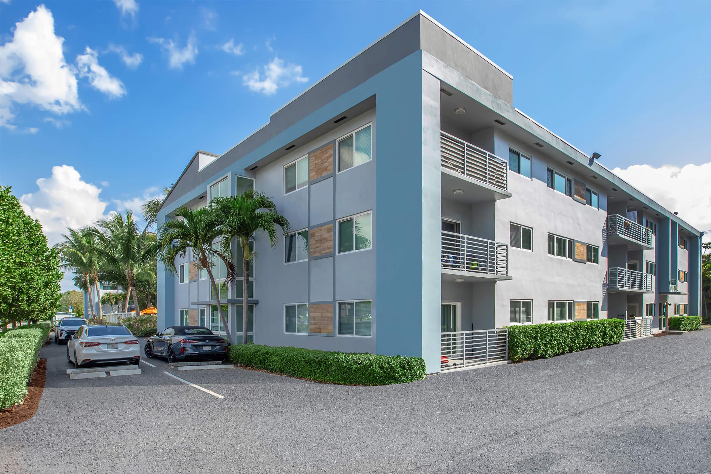 A modern, light blue apartment building with multiple balconies surrounded by lush palm trees. The building features a neatly paved parking area with two parked cars. The sky is bright with a few scattered clouds, creating a pleasant atmosphere.