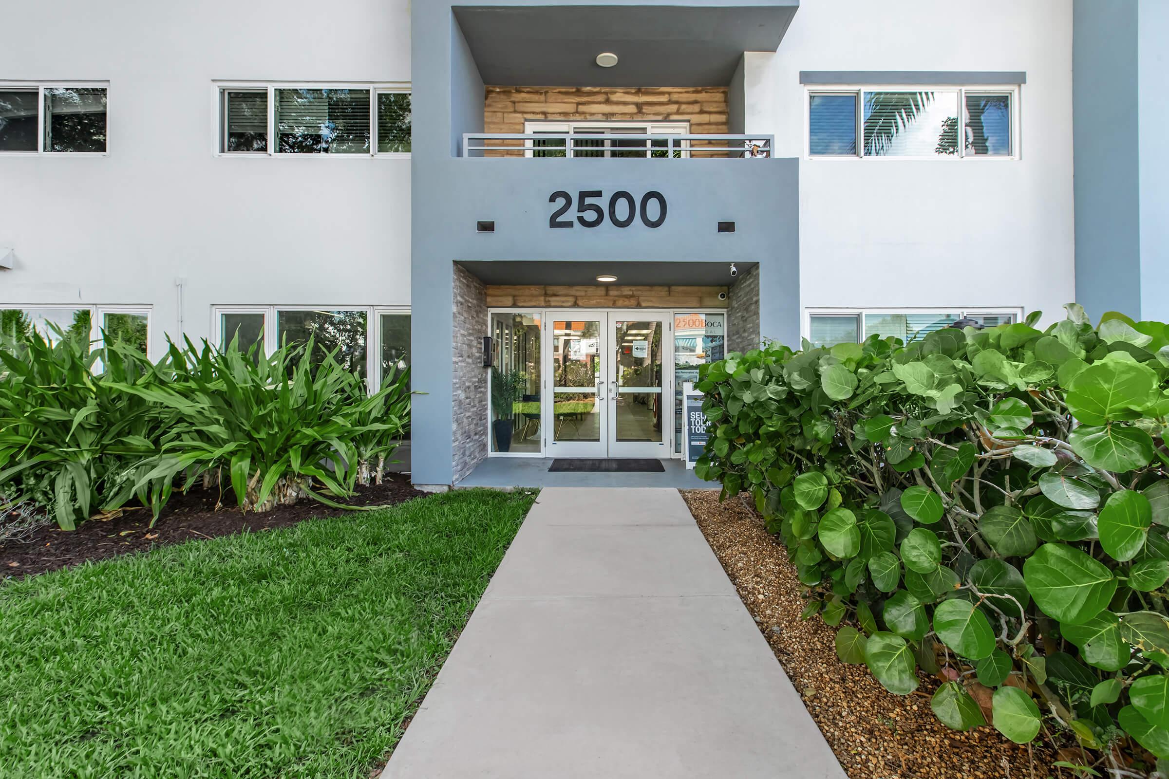 Exterior view of a modern building entrance featuring large glass doors with the number "2500" displayed above. The pathway is lined with green grass and well-maintained shrubs, leading to the entrance. Large windows provide a glimpse of the interior space.