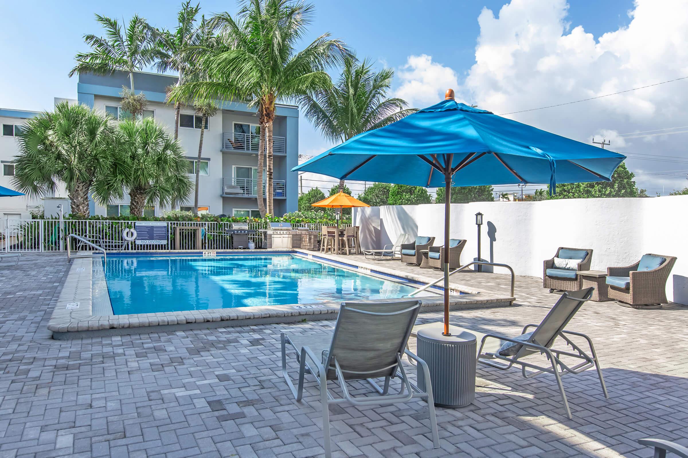 A serene swimming pool surrounded by palm trees and lounge chairs. An umbrella shades the seating area, with additional outdoor furniture nearby. The pool area features a clean, modern design and is bathed in sunlight against a backdrop of blue skies and fluffy clouds.