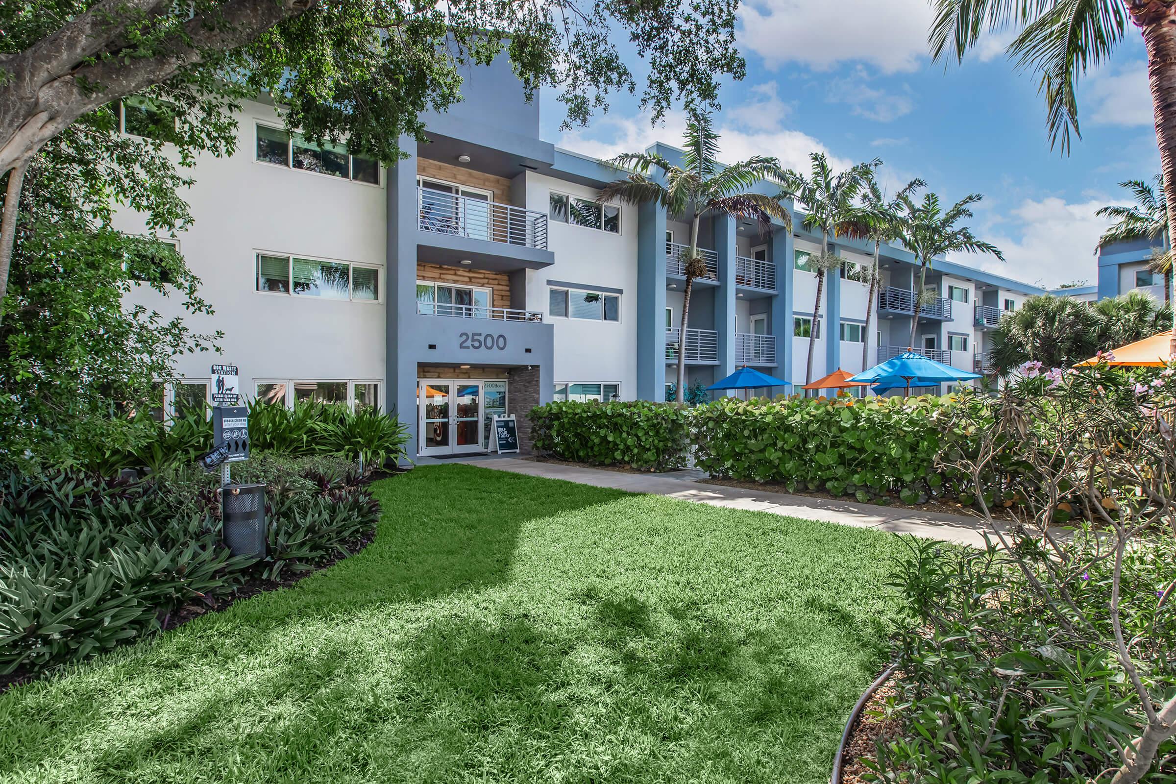 Contemporary apartment building with a lush green lawn and landscaping. The entrance features large windows and a blue awning. In the foreground, there are tropical plants and vibrant orange and blue umbrellas, indicating an inviting outdoor area. Clear blue sky with a few clouds can be seen above.