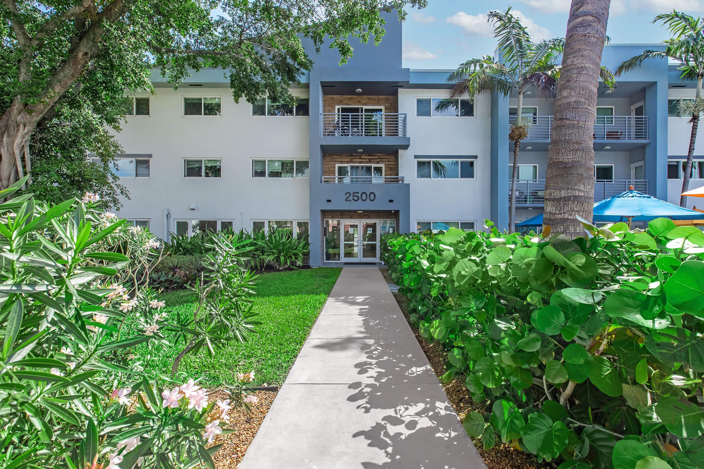 A walkway leading to a modern apartment building with the number "2500" displayed prominently. The building features multiple balconies, surrounded by lush greenery and palm trees. Bright blue umbrellas are visible in the background, creating a welcoming outdoor atmosphere.