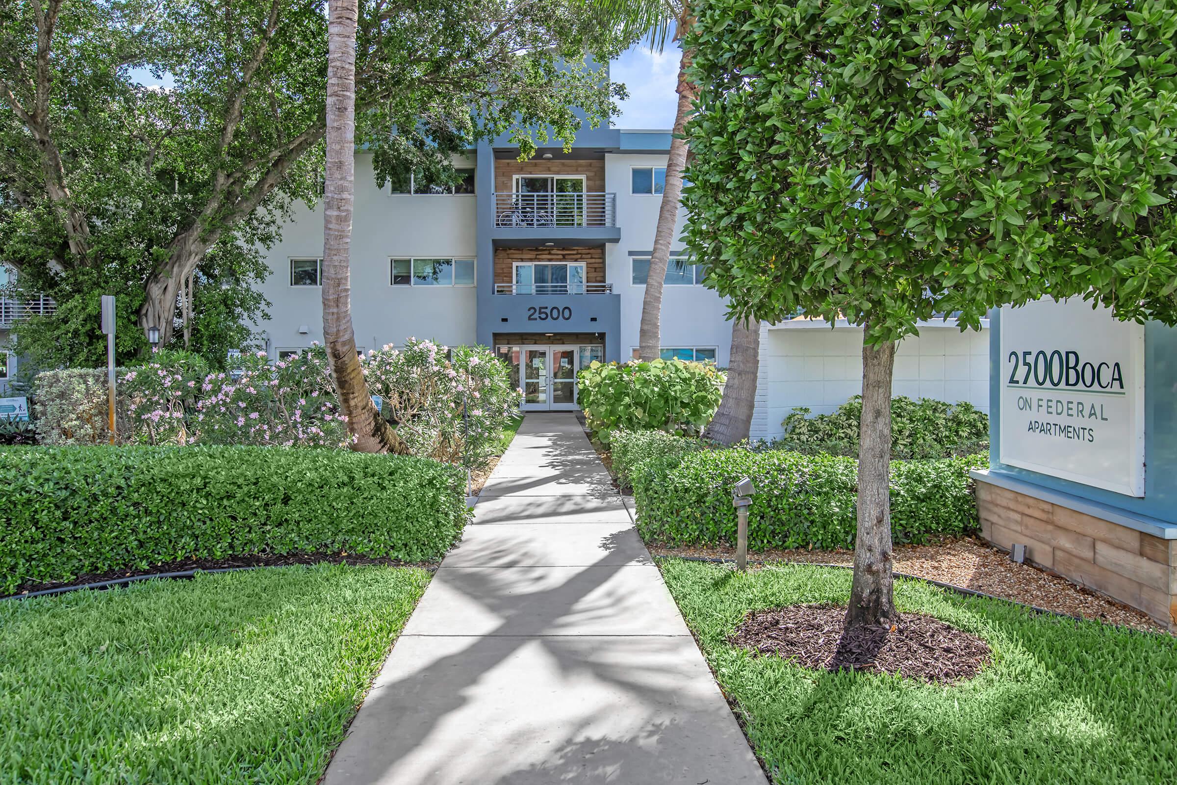 Pathway leading to the entrance of 2500 Boca on Federal Apartments, lined with lush green grass and shrubs. The building features a modern design with balconies and palm trees, creating a welcoming atmosphere. Flowering plants add a pop of color to the landscaped area.