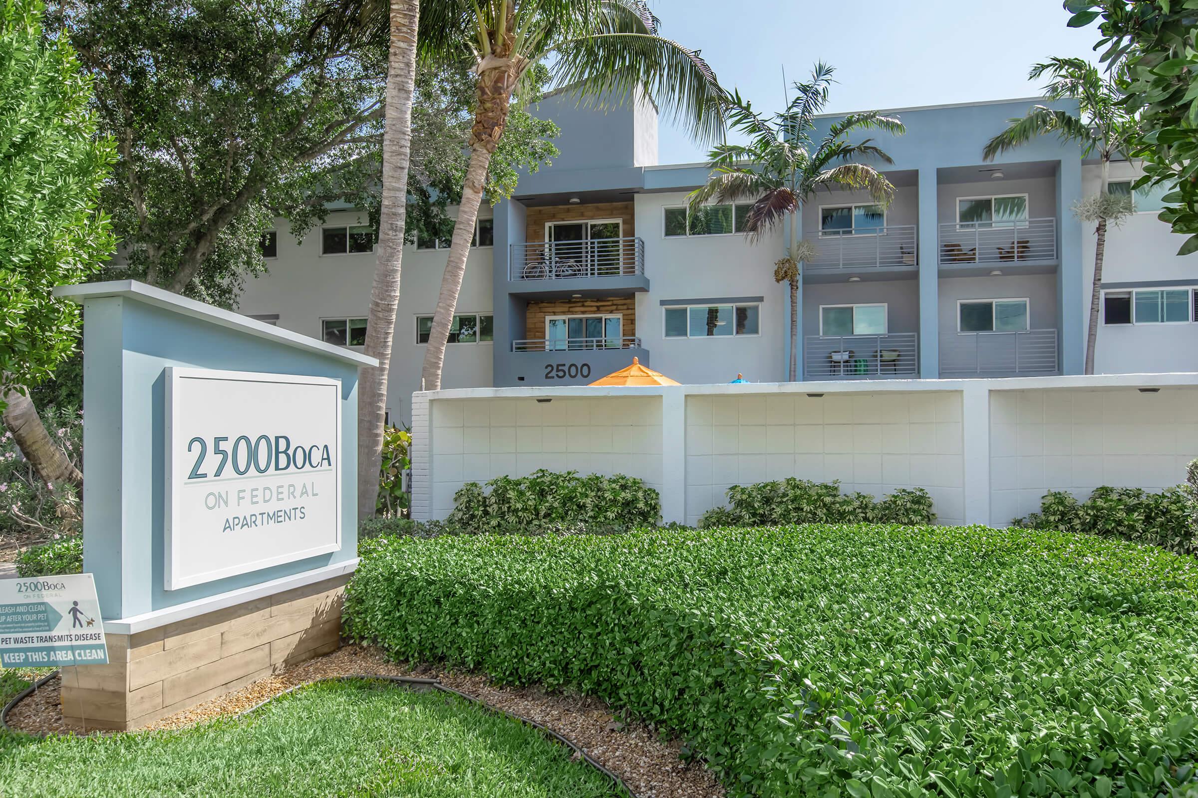 A modern apartment complex named "2500 Boca on Federal Apartments," featuring a welcoming sign, lush green landscaping, and palm trees. The building has multiple balconies and is situated in a sunny, tropical environment.